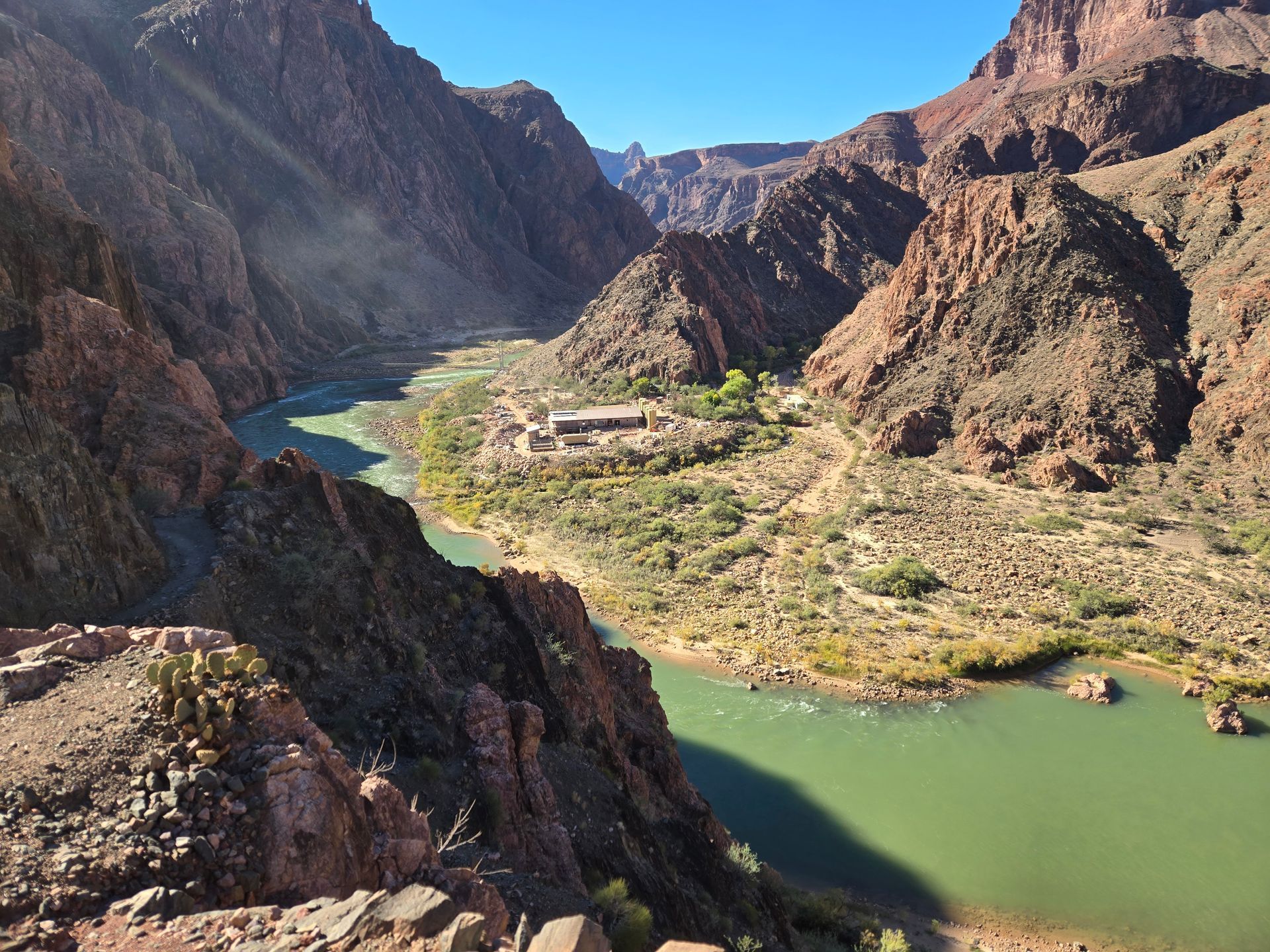 View of the Grand Canyon's river with a lodge surrounded by green vegetation, canyon walls in sunlight.
