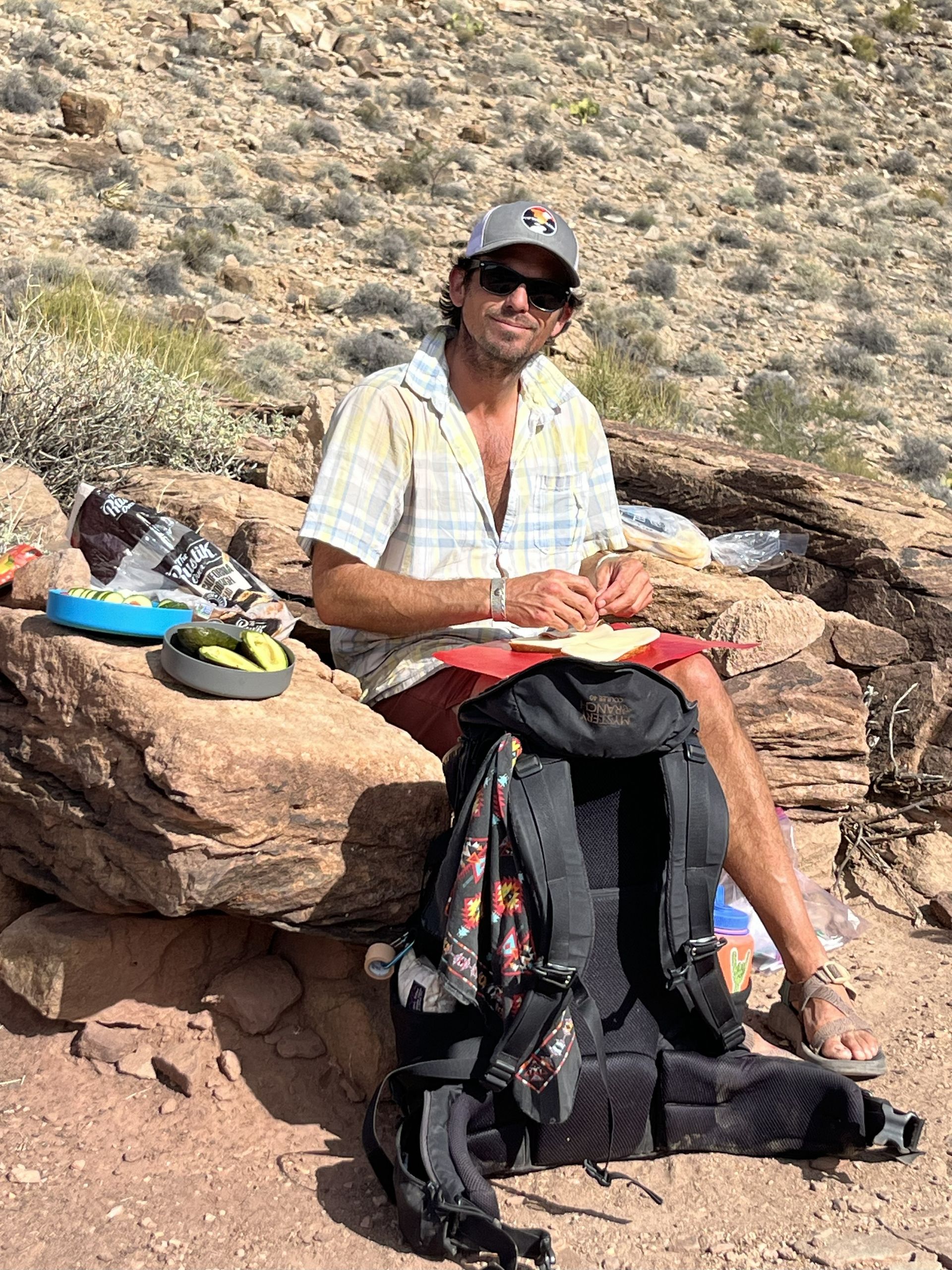 Man in hat, sunglasses, shirt, and shorts sits on rocks, backpack next to him. Desert background.