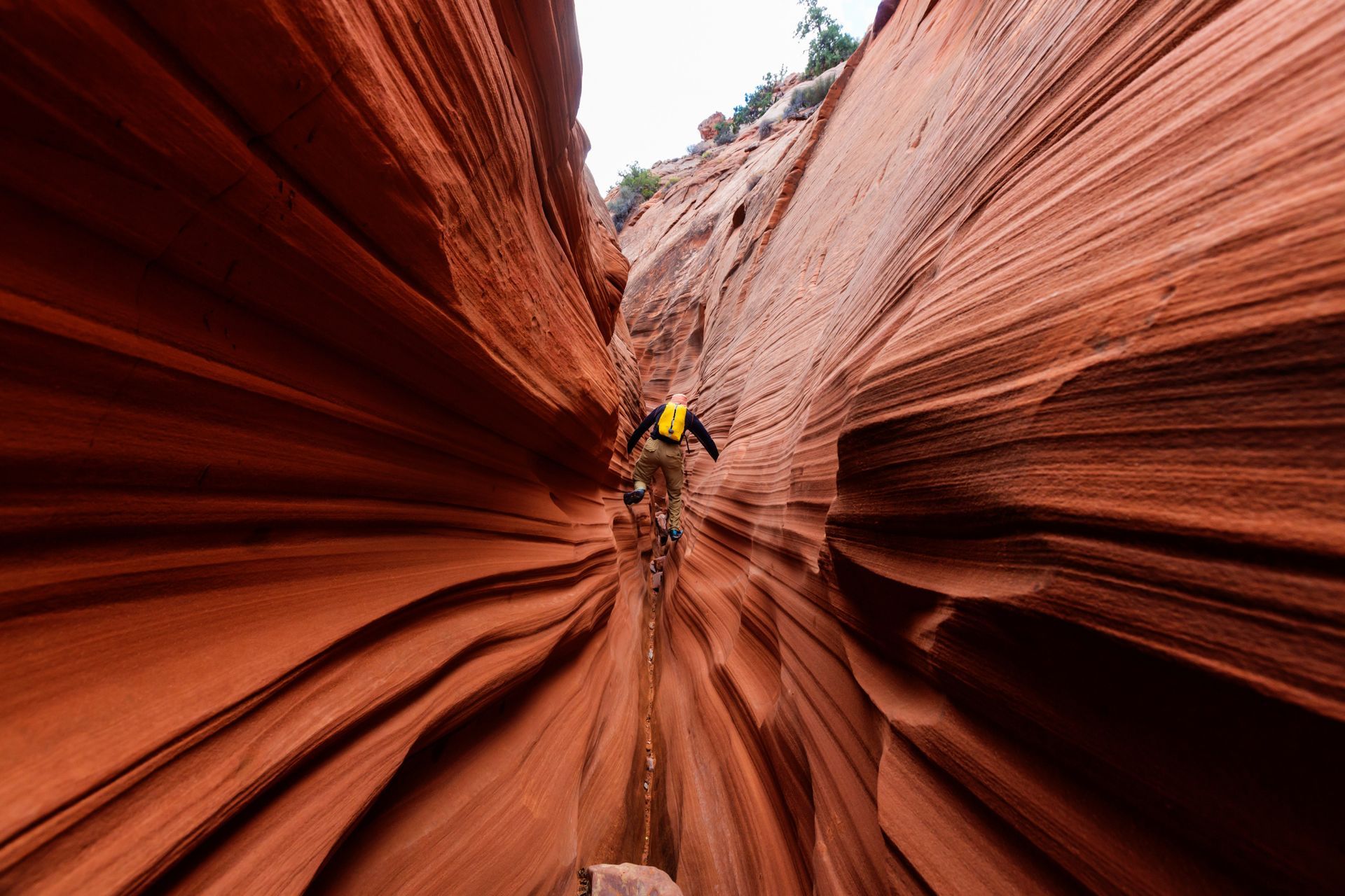 A person in a yellow jacket hikes through a narrow, winding slot canyon with textured, reddish-orange sandstone walls.
