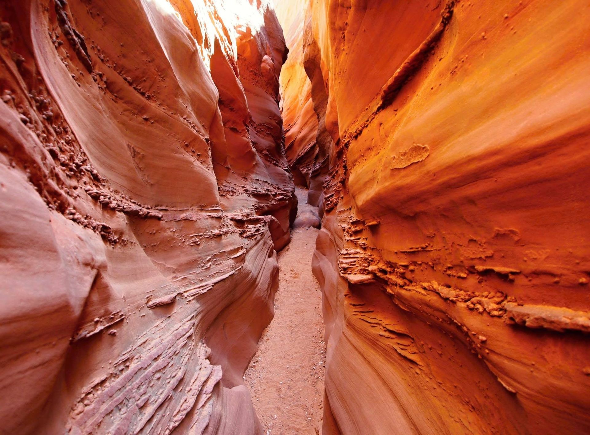 A narrow, winding desert slot canyon with tall, smooth, vibrant orange and red sandstone walls and a sandy floor.