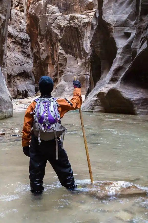 A person with a backpack is standing in a river holding a stick.