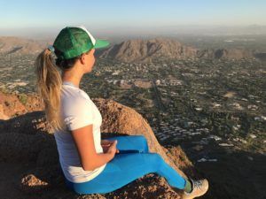 A woman is sitting on top of a mountain looking out over a city.