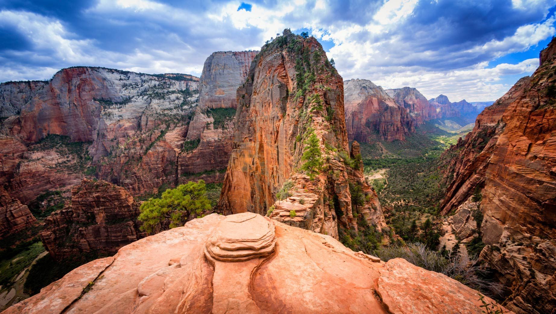 Red rock mountain ridge with a narrow trail, lush green valley, and cloudy sky.