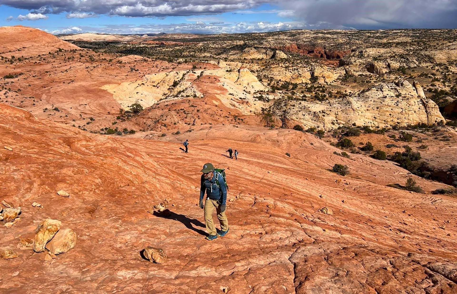 People with backpacks are hiking down a trail next to a cliff.