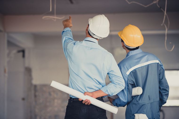 Zwei Bauarbeiter mit Schutzhelmen begutachten einen Deckenplan auf einer Baustelle für Innenrenovierungen.