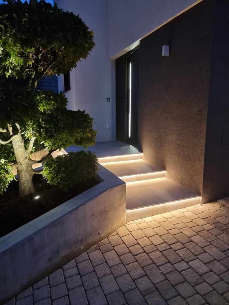Beleuchtete Außentreppe neben einem modernen Haus und einem gestutzten Baum in der Abenddämmerung