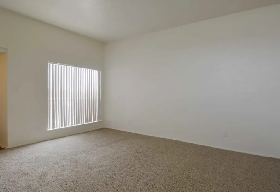Empty living room with beige walls, plush carpet, and a window with vertical blinds at Brookview, offering apartments for rent near Music City Mall in Odessa, TX.