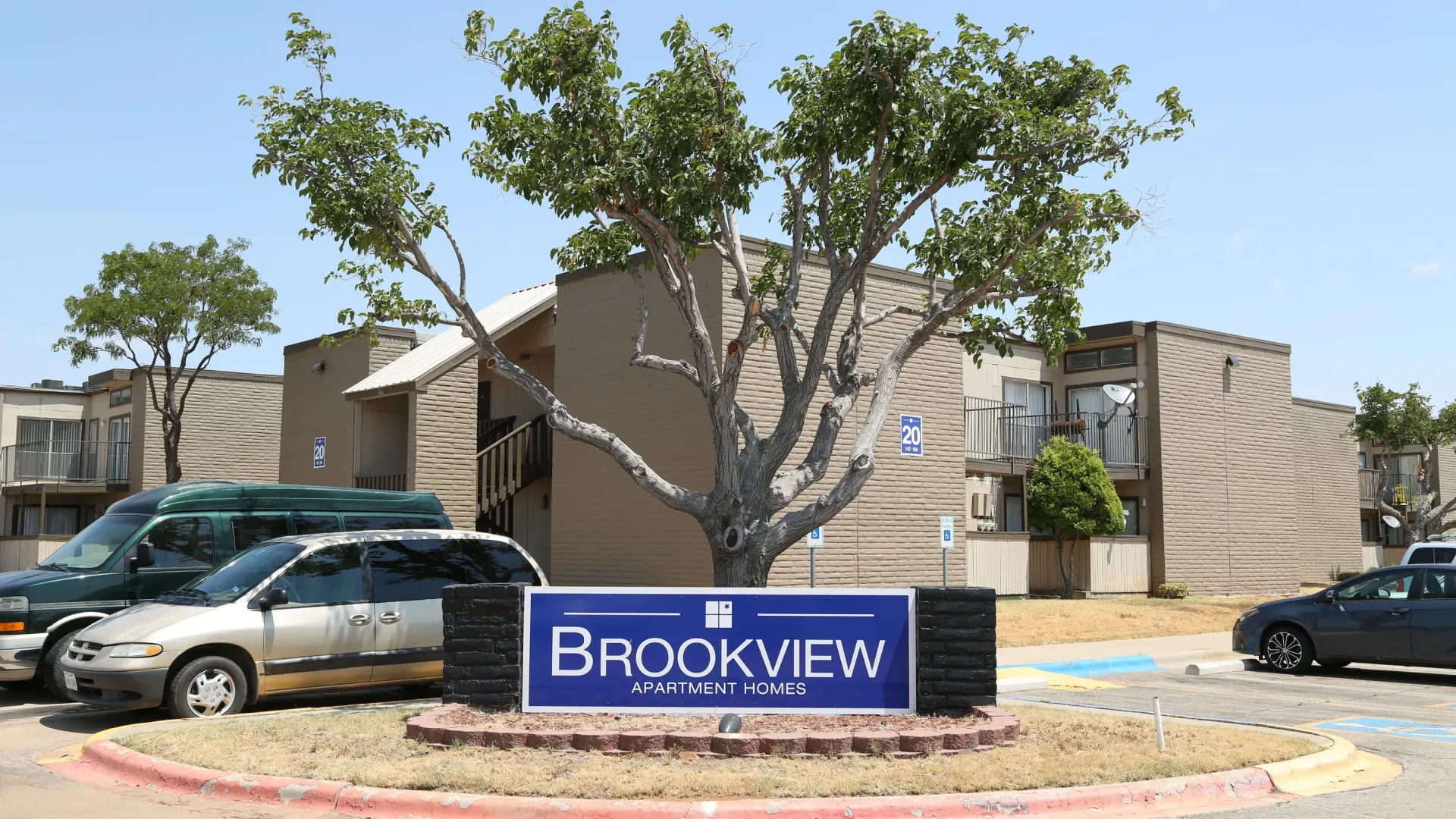 Brookview Apartments sign in front of a tan two-story building; cars and a van parked in front.