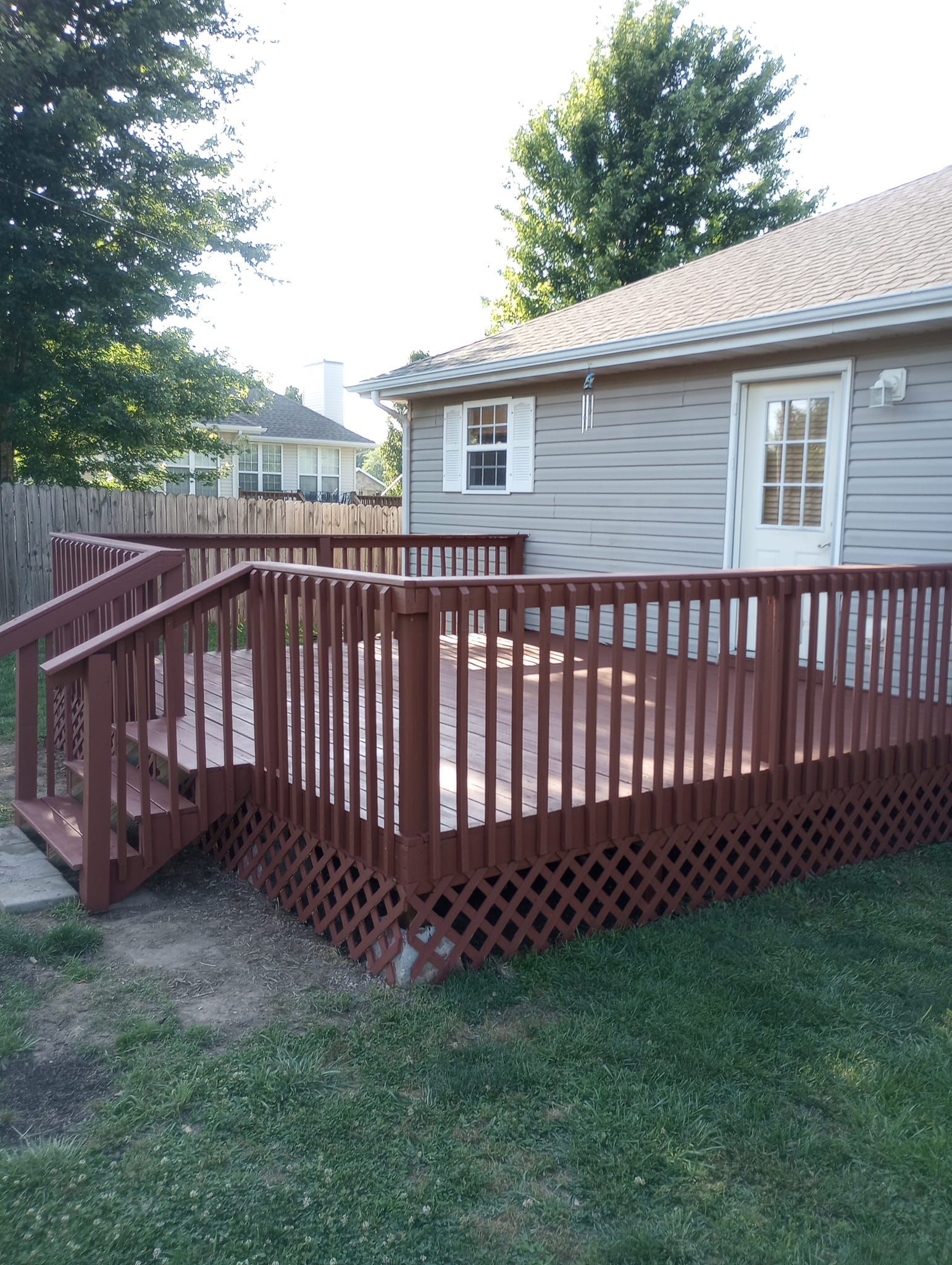 Wooden deck attached to a house with a door and window; brown railing and lattice, backyard setting.