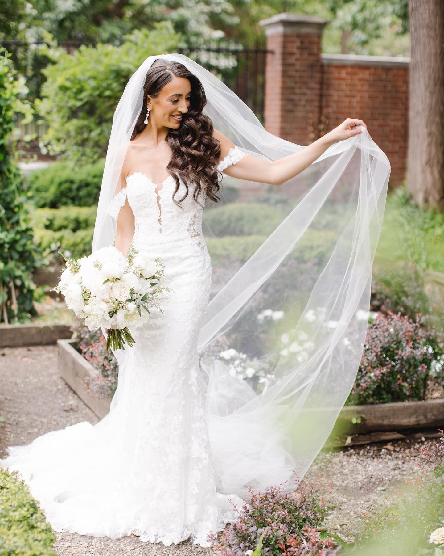 Bride in a white gown holding a bouquet and veil outdoors in a garden setting