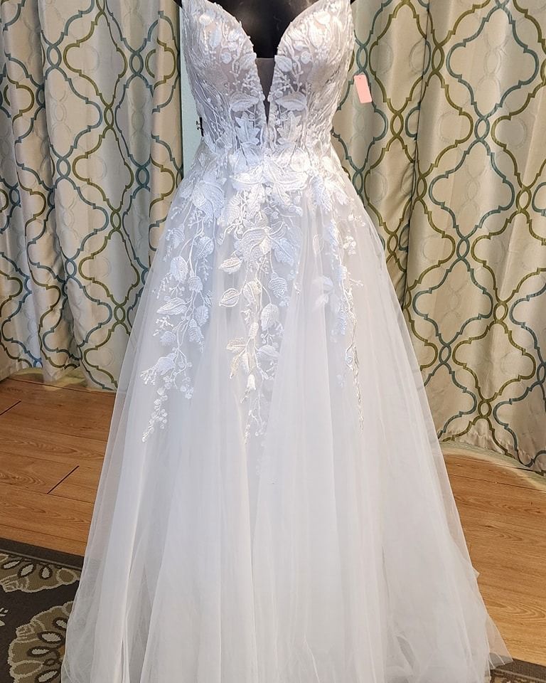 White lace strapless wedding gown with floral detailing on a mannequin against patterned curtains.