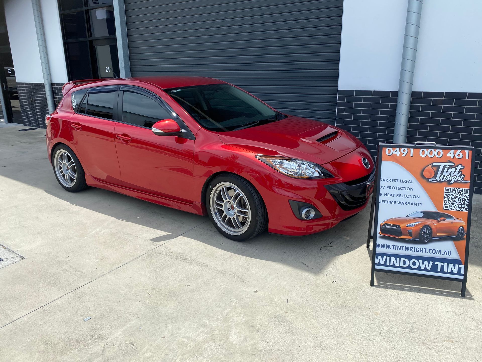 A red car is parked in front of a building next to a sign.