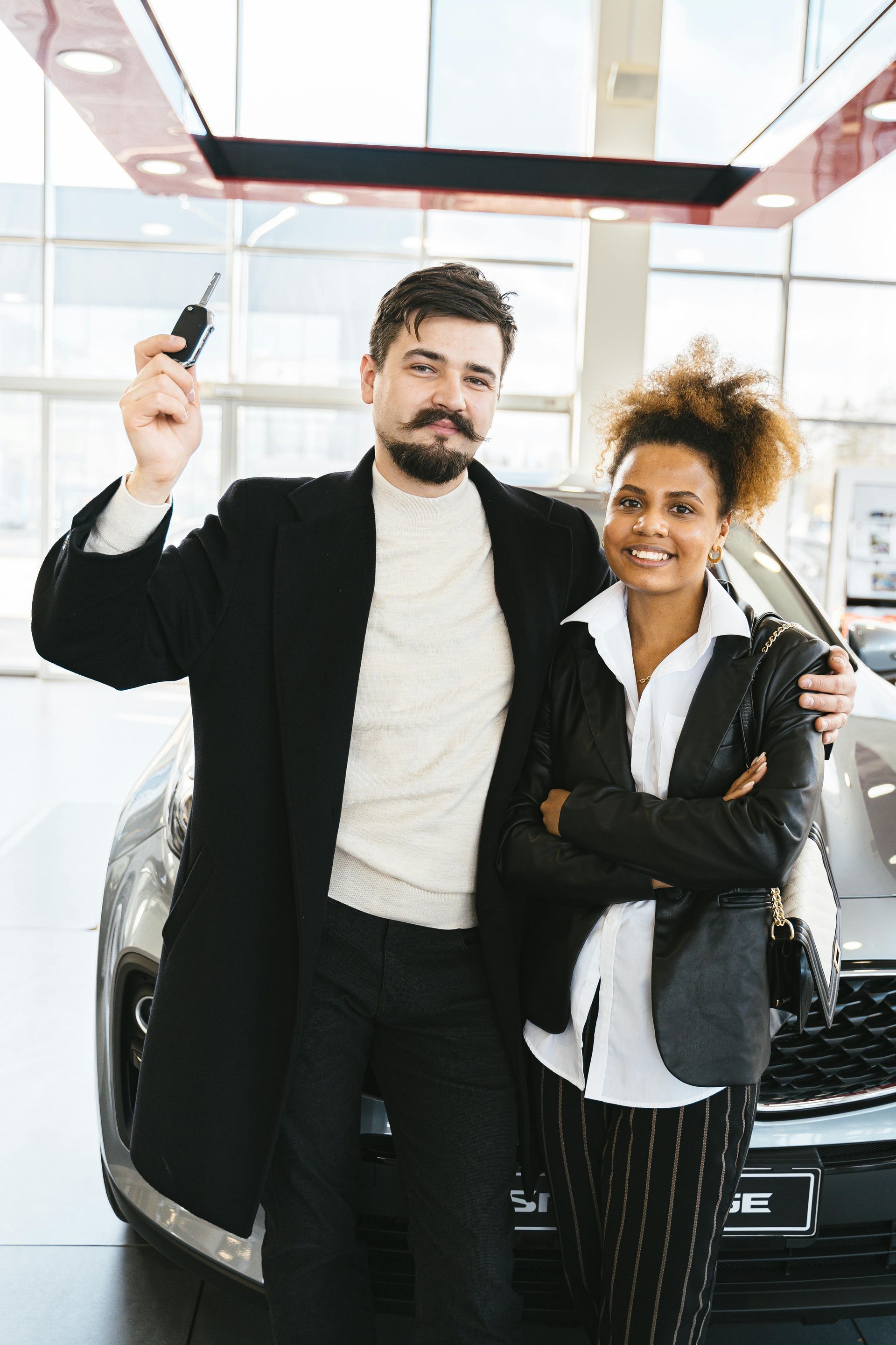 A man and a woman are standing next to a car in a showroom . the man is holding a car key.