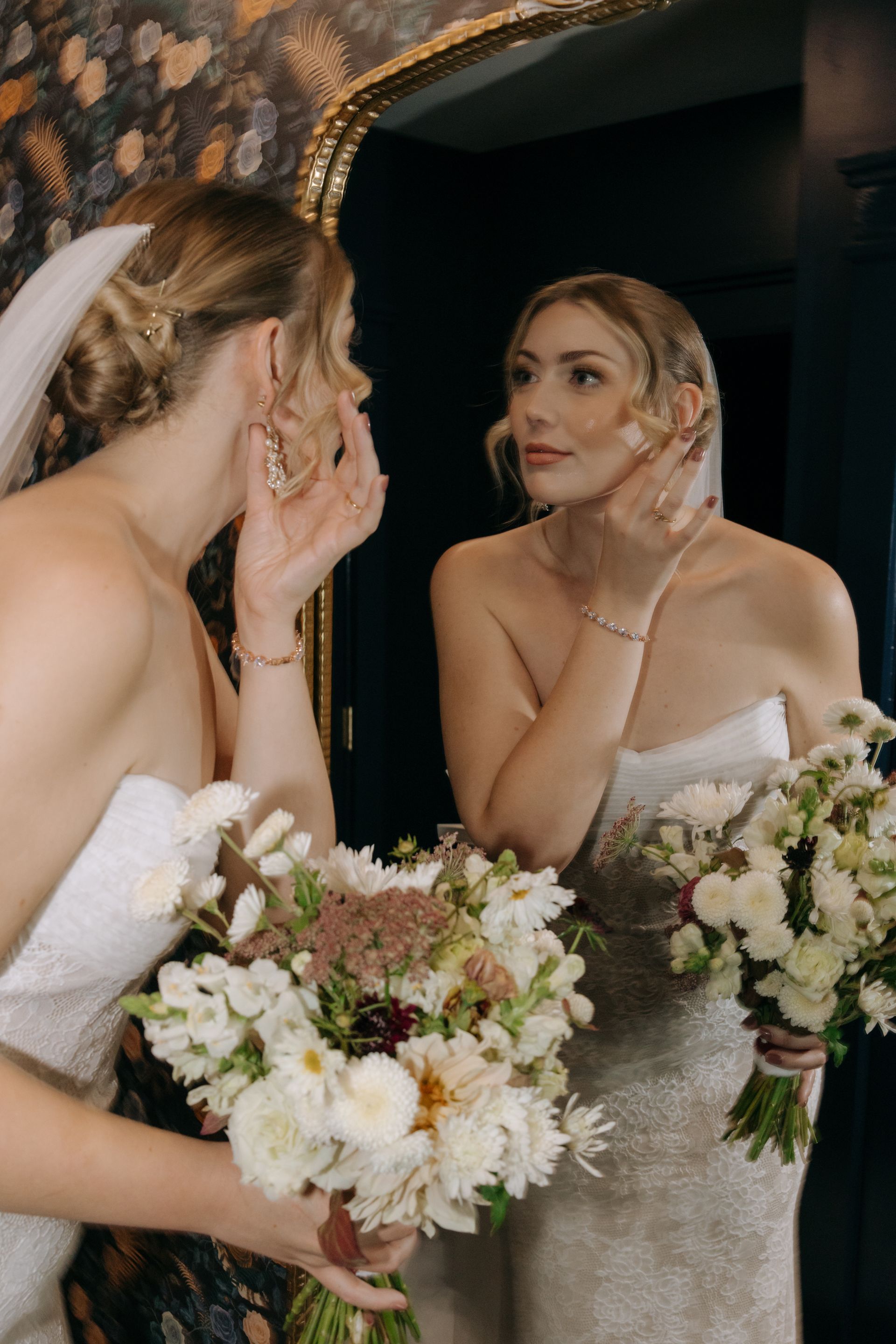 Bride adjusting earring in mirror, holding white floral bouquet in a dimly lit doorway.