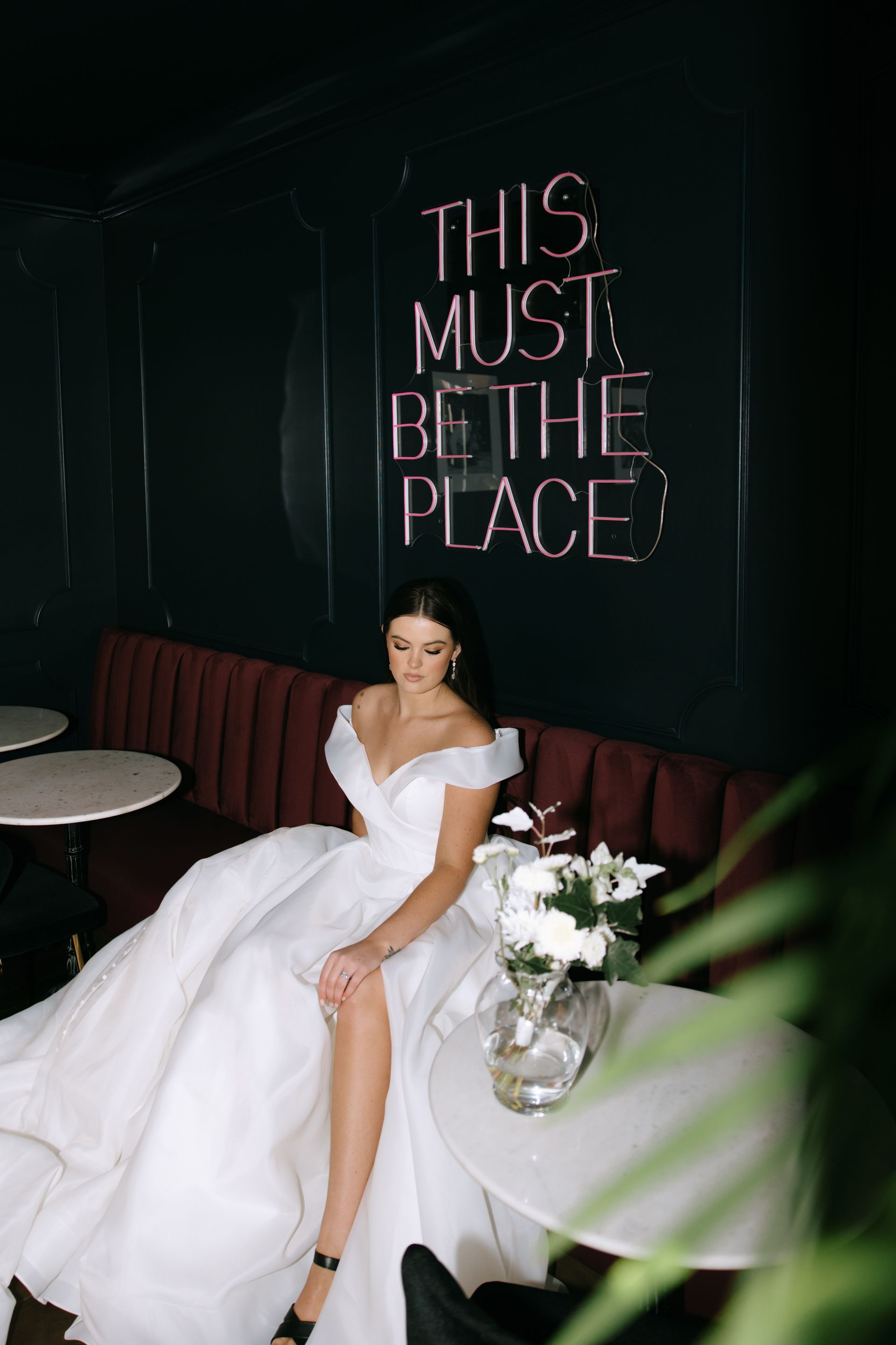 Woman in white dress sitting on a couch beneath a neon sign that reads “THIS MUST BE THE PLACE”