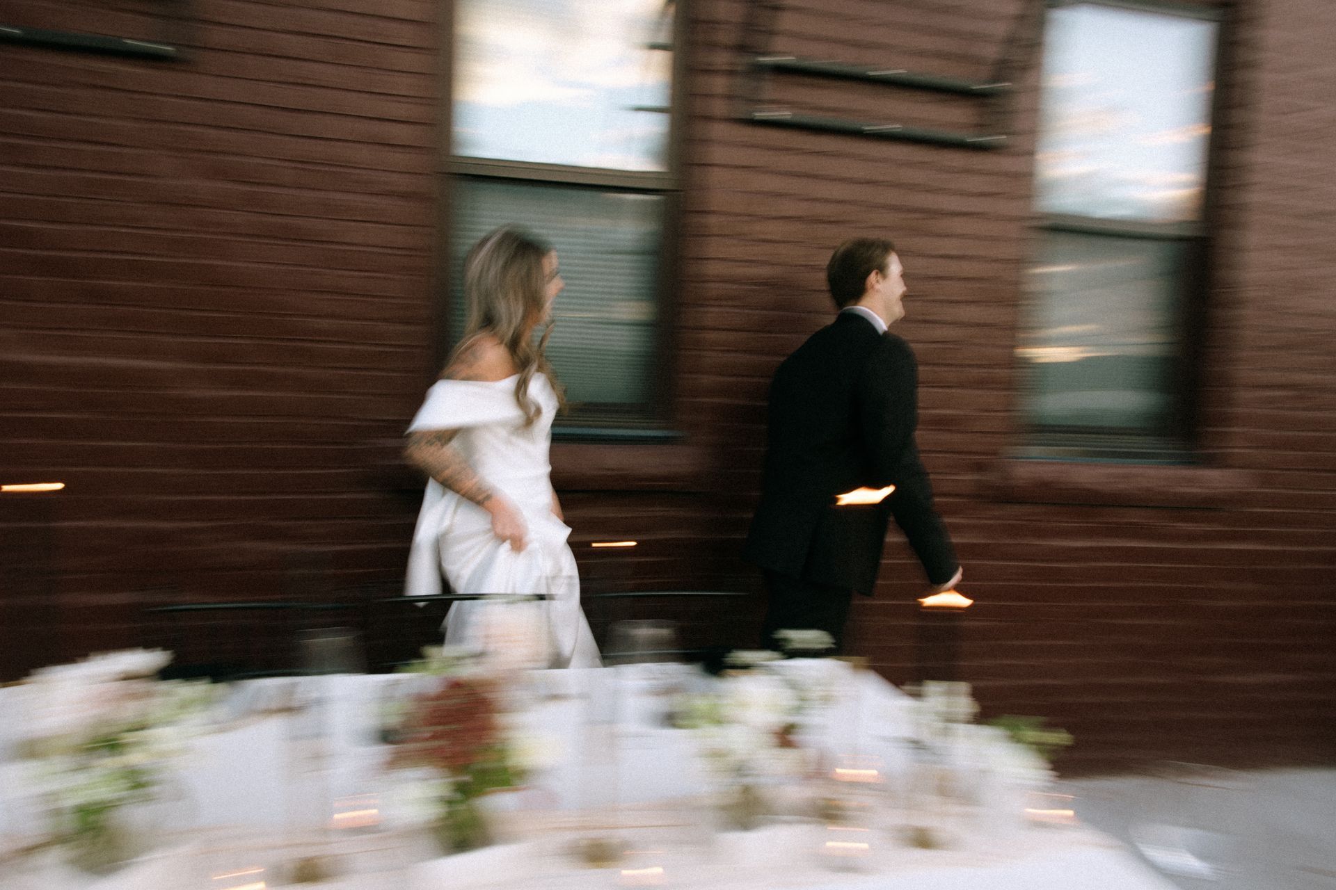 Bride in white dress and groom in black suit walking past a brick building outdoors.
