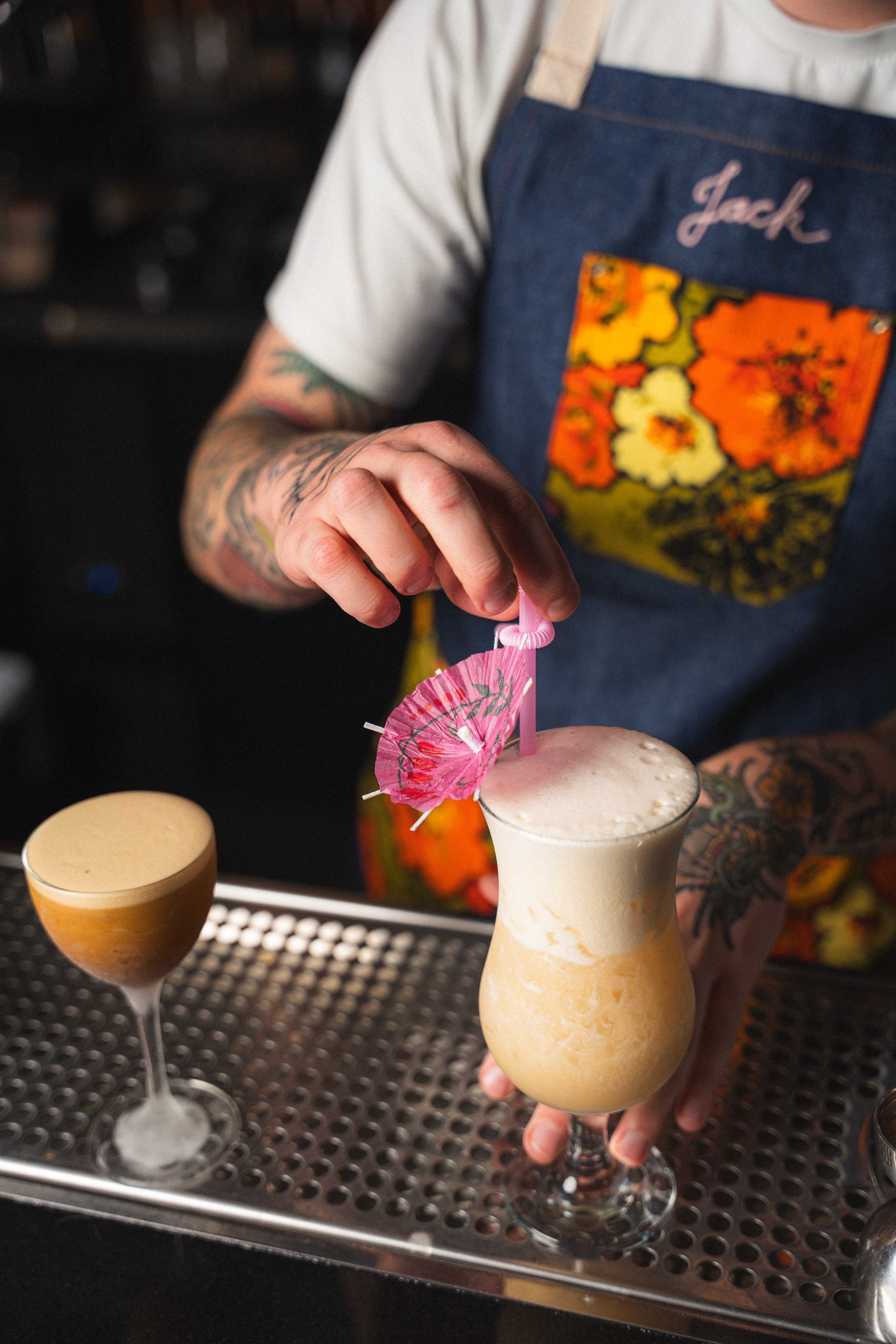 Bartender pouring a pink-topped cocktail into a stemmed glass on a bar counter