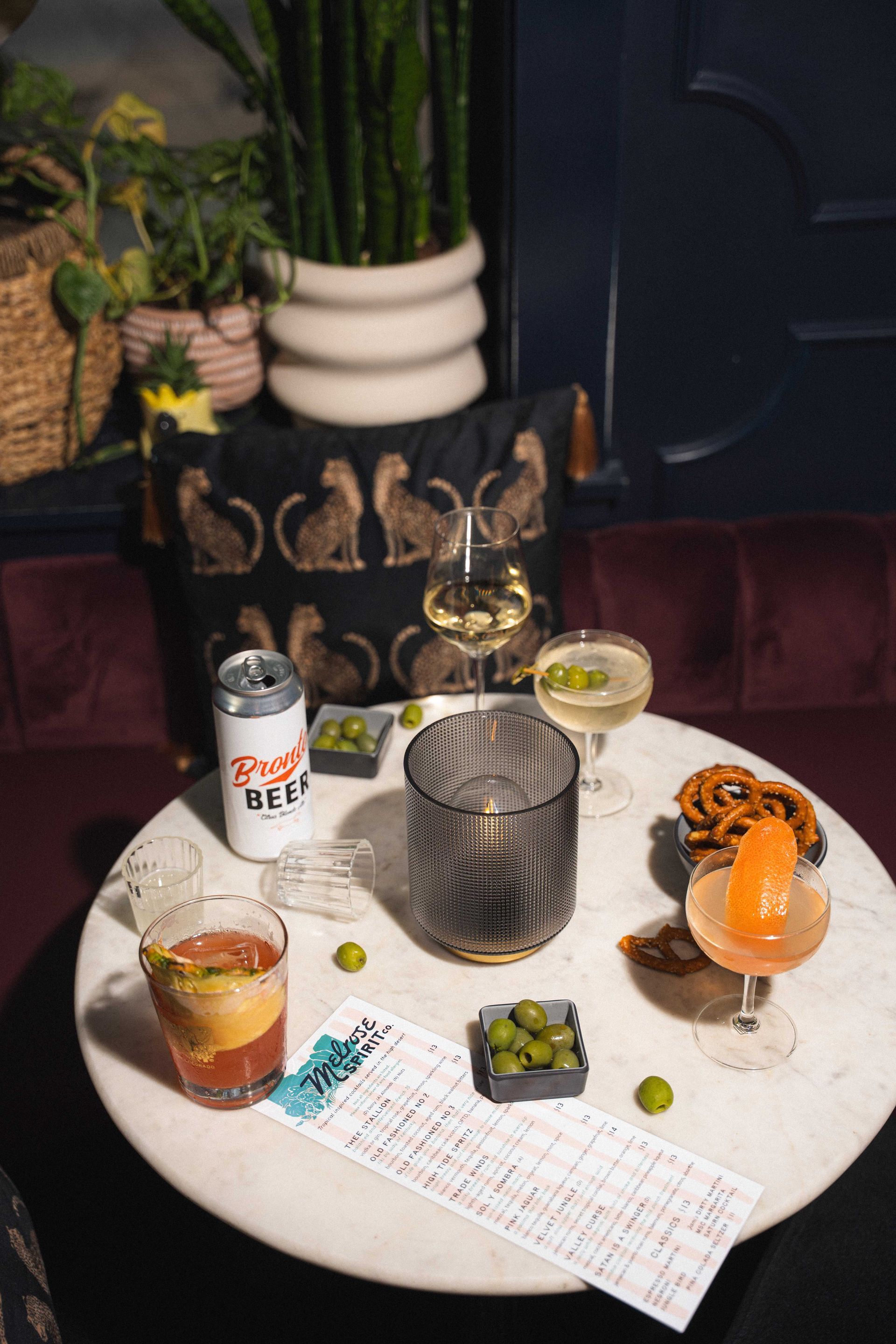 Cocktail on a white table with menu, snacks, and a can in a dim lounge setting
