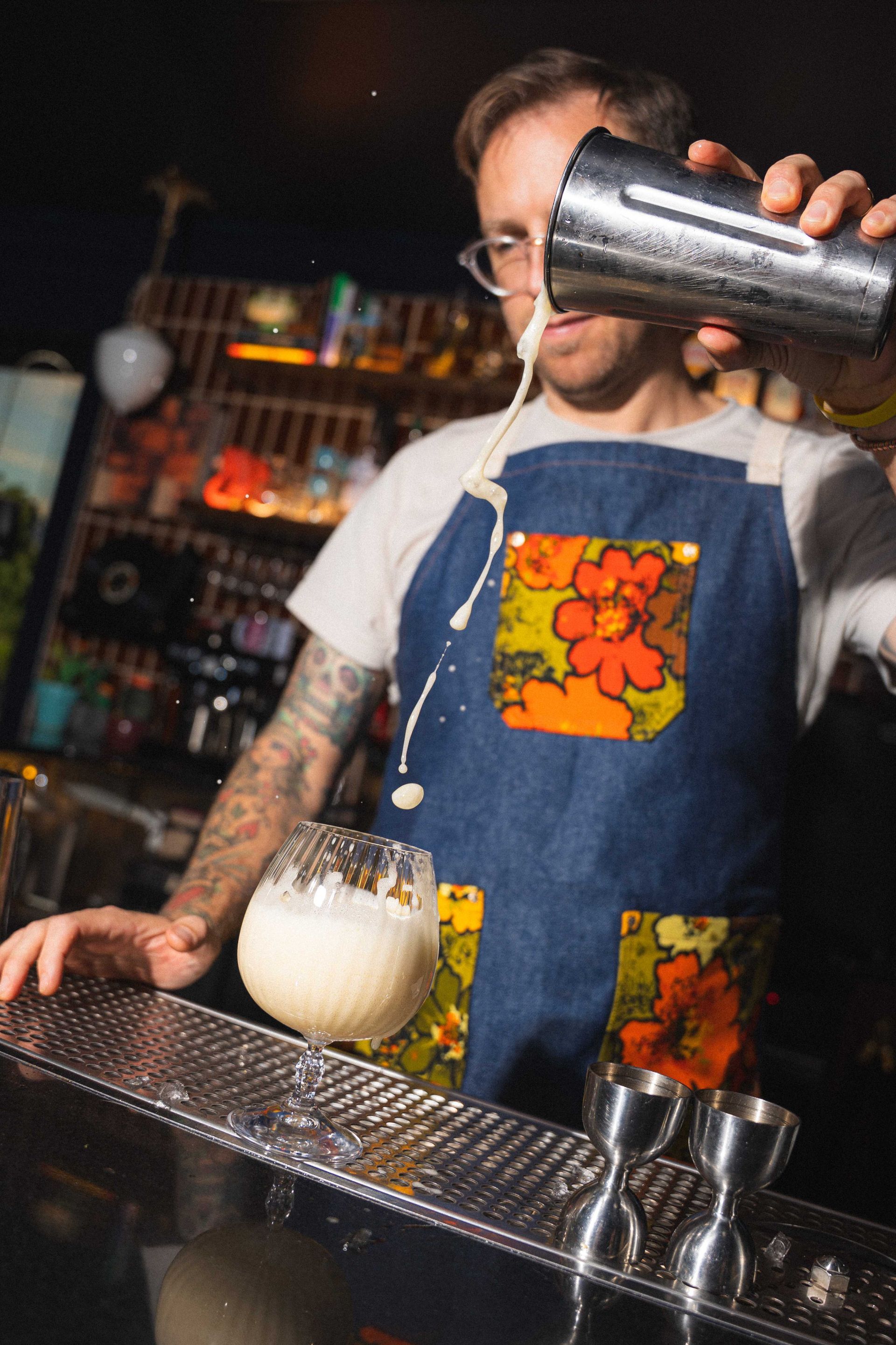 Bartender pouring a drink into a glass at a bar, wearing a blue apron with orange floral patches.