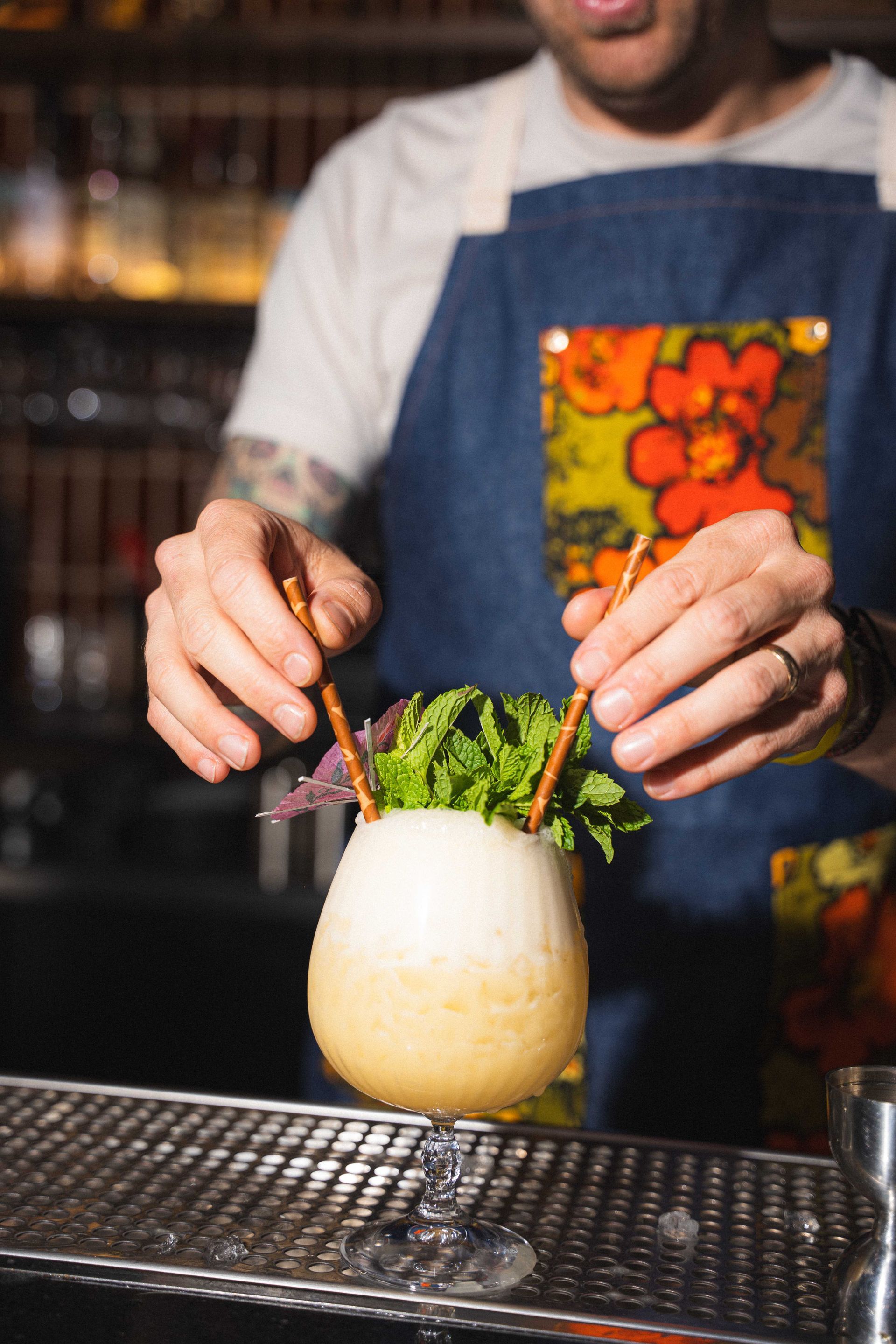 Bartender garnishes a frothy cocktail with herbs behind a bar.