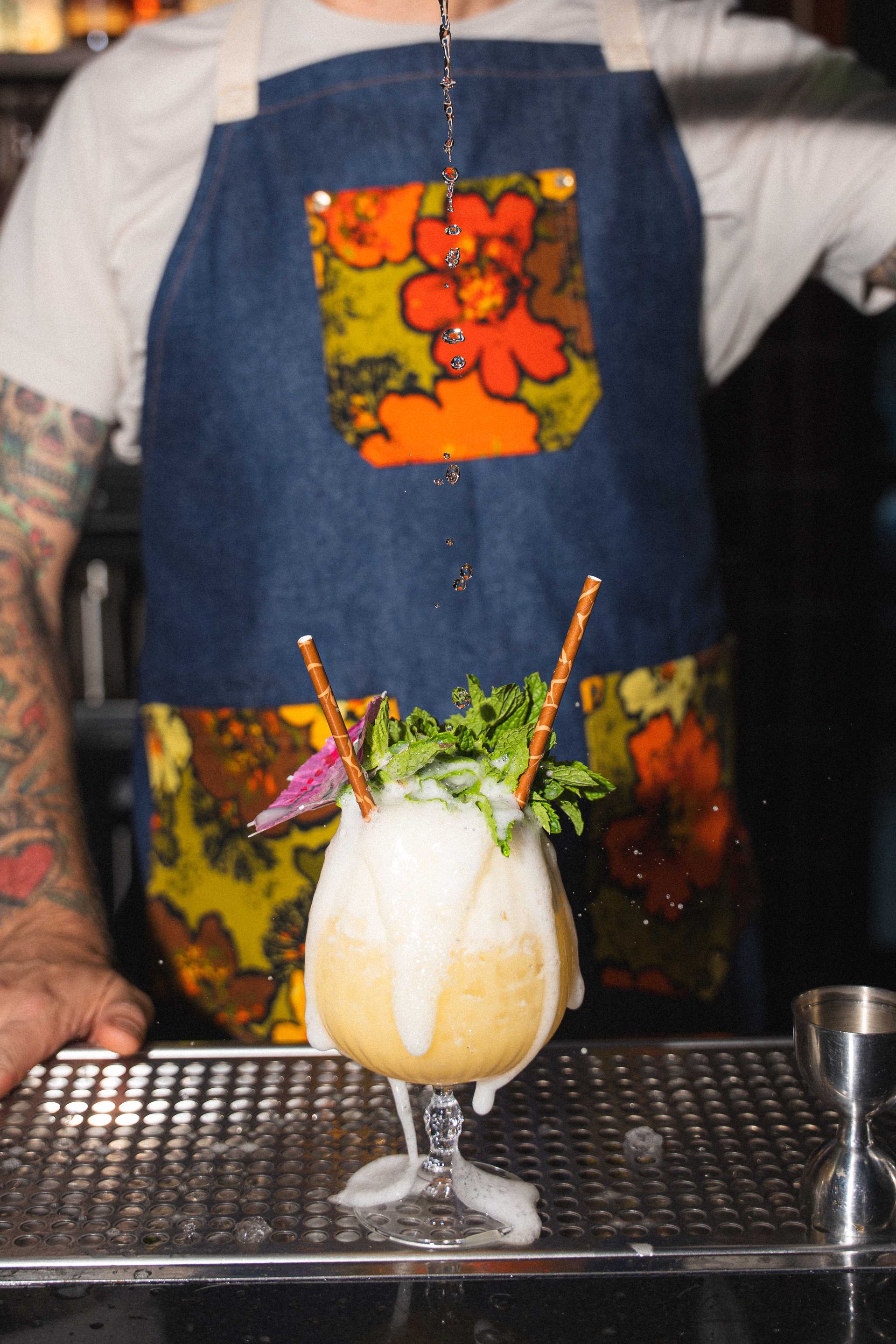 Bartender behind a bar holds a frosty cocktail topped with herbs and garnish