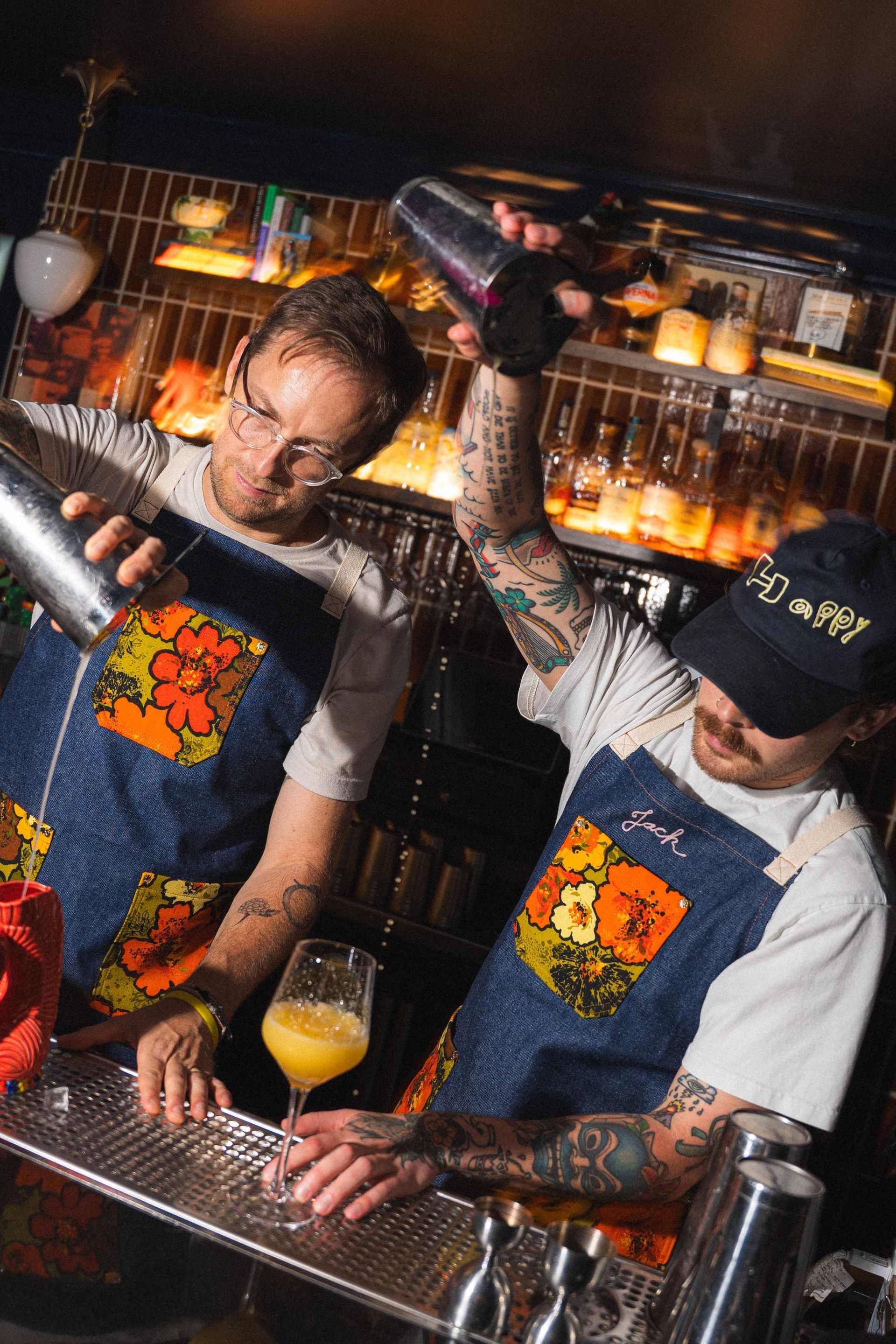 Two bartenders in aprons shake cocktails behind a lit bar, surrounded by bottles and glassware.