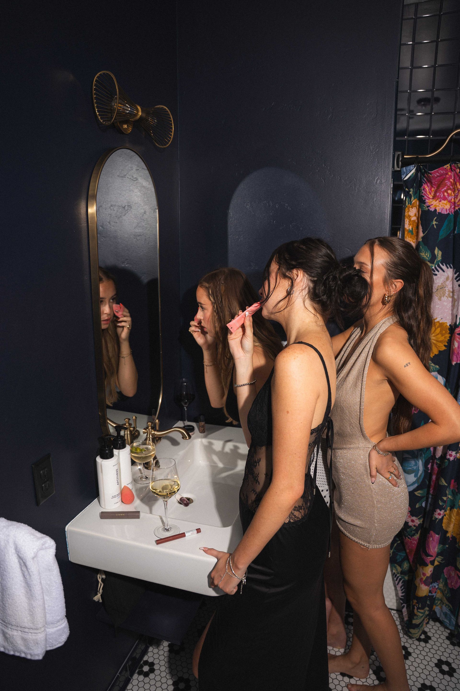 Three people standing at a bathroom sink, looking in the mirror in a dimly lit room.
