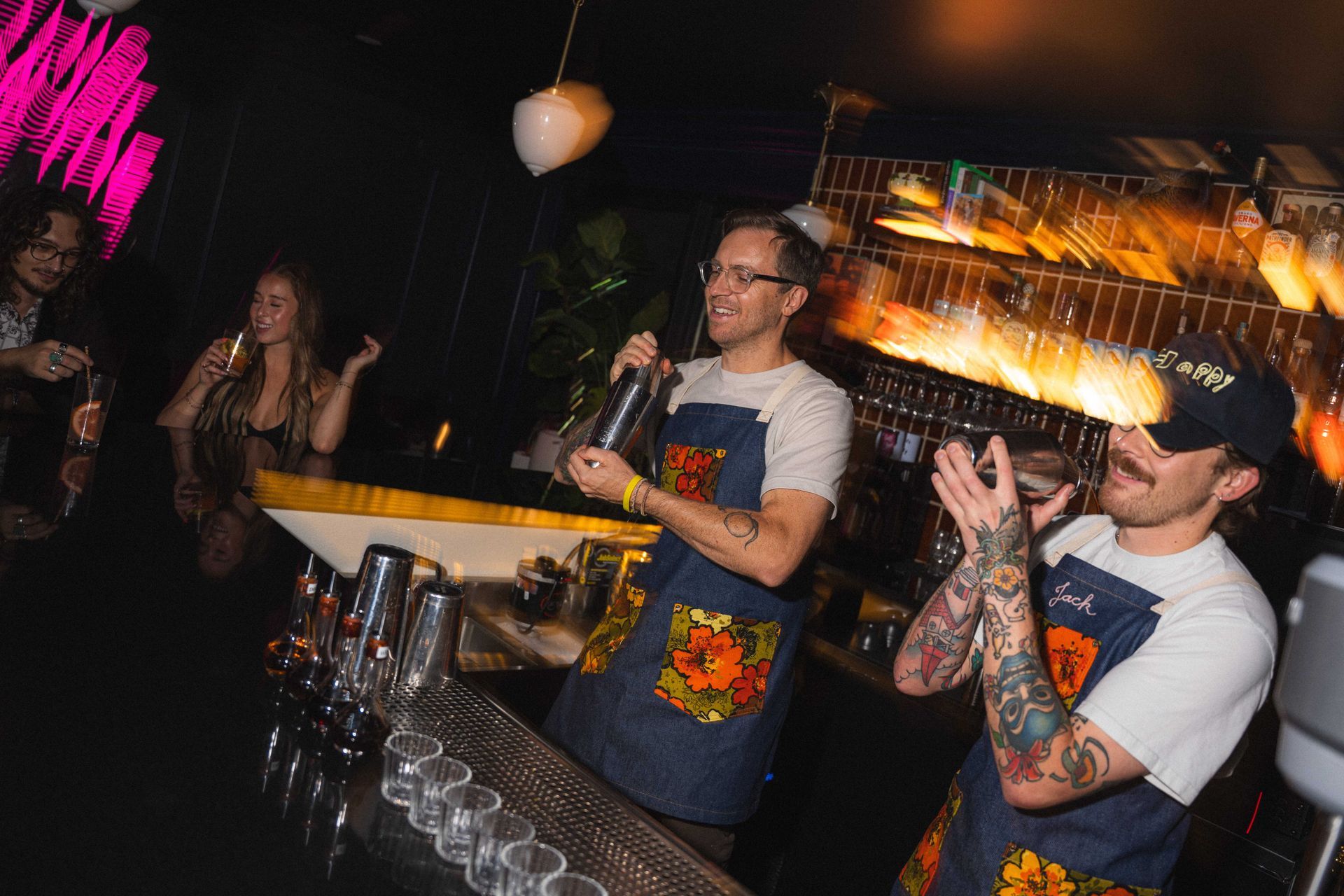 Bartenders serving drinks at a lively nightclub bar with glowing shelves and rows of glasses