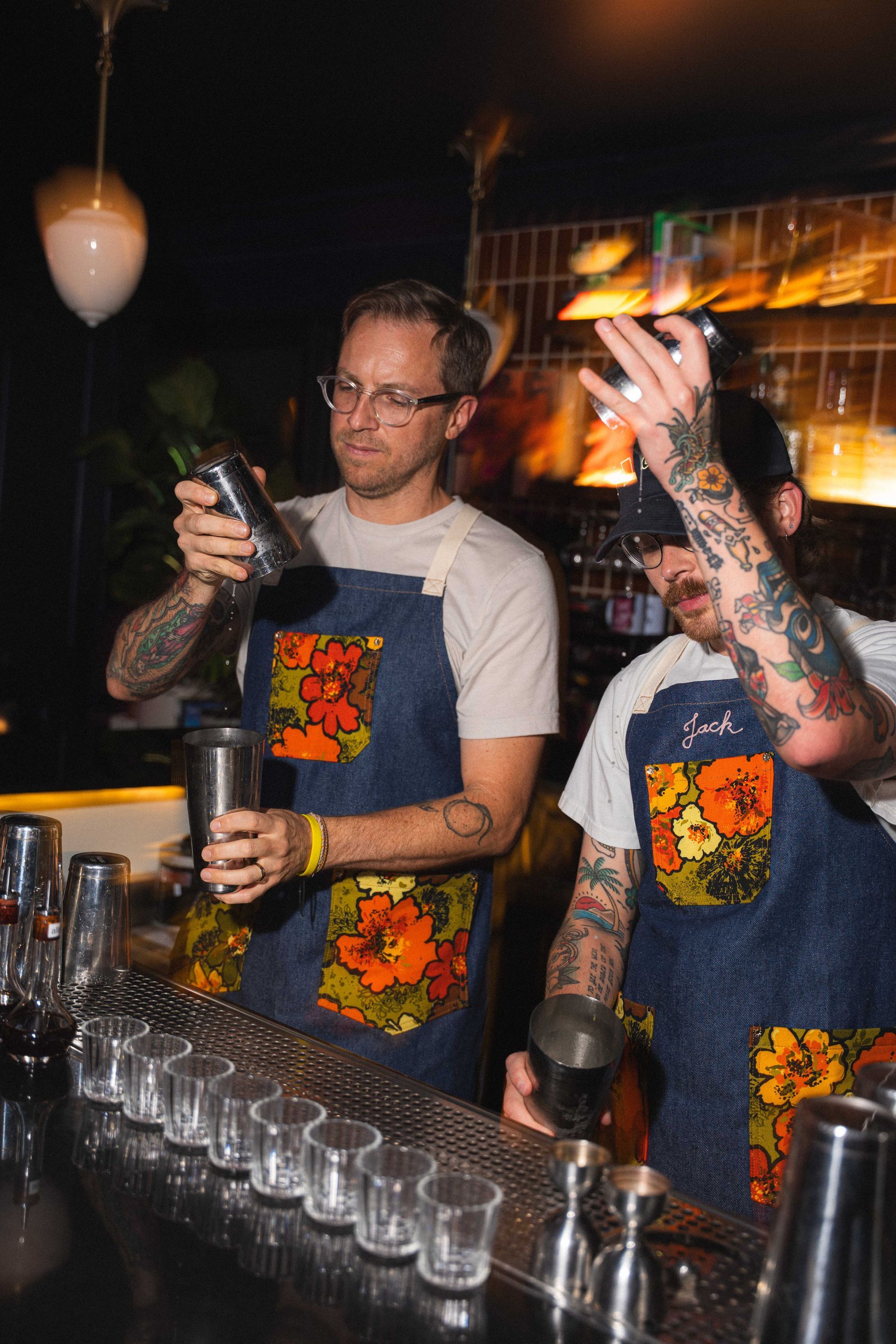 Bartenders in matching aprons mixing drinks behind a bar with rows of shot glasses