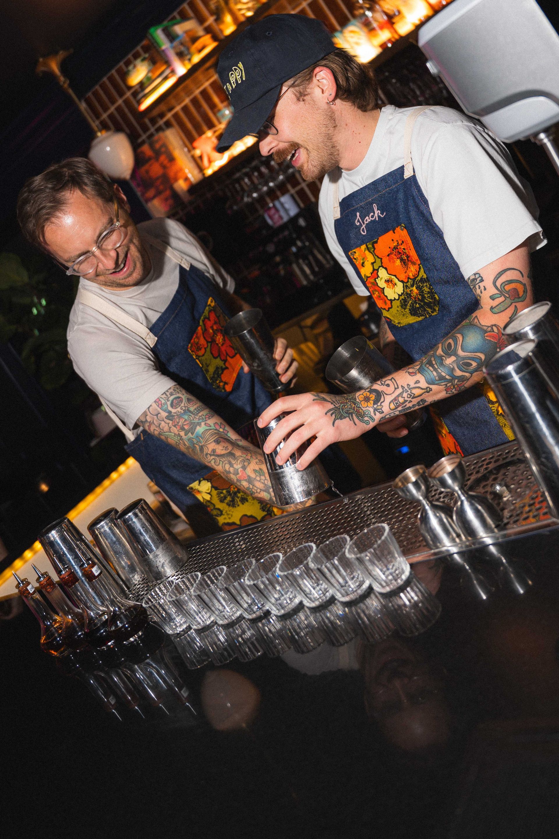 Two bartenders pouring drinks behind a bar lined with rows of glasses and bottles