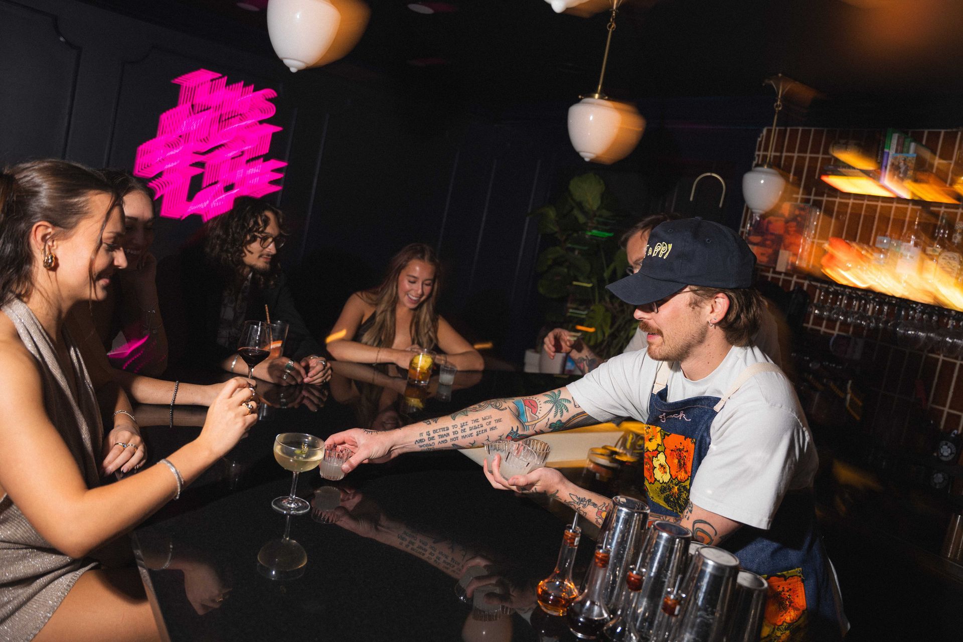 People chatting and toasting at a dimly lit bar with drinks and neon pink wall art