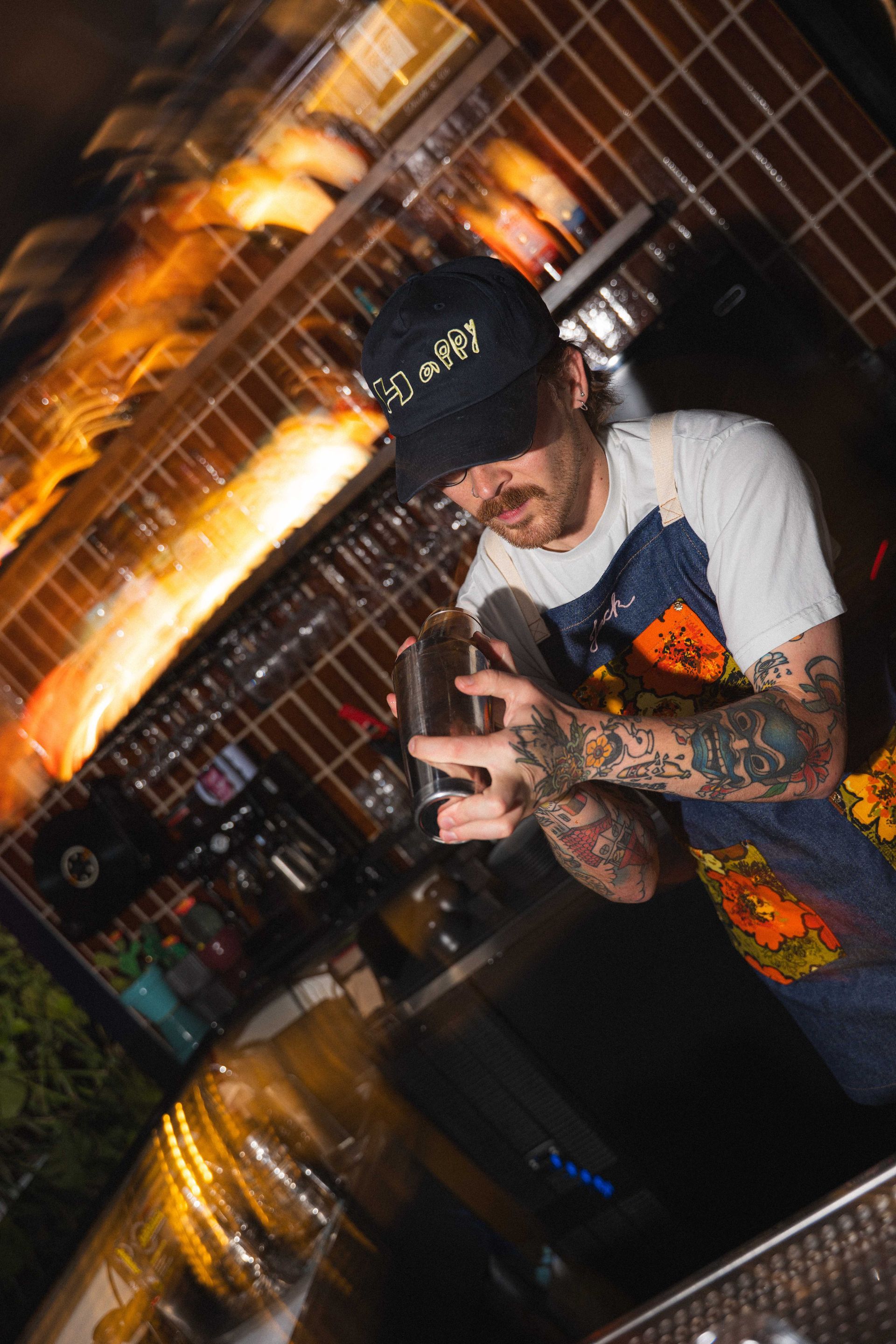 Tattooed person in a black cap flashing a peace sign behind a bar with warm orange lights