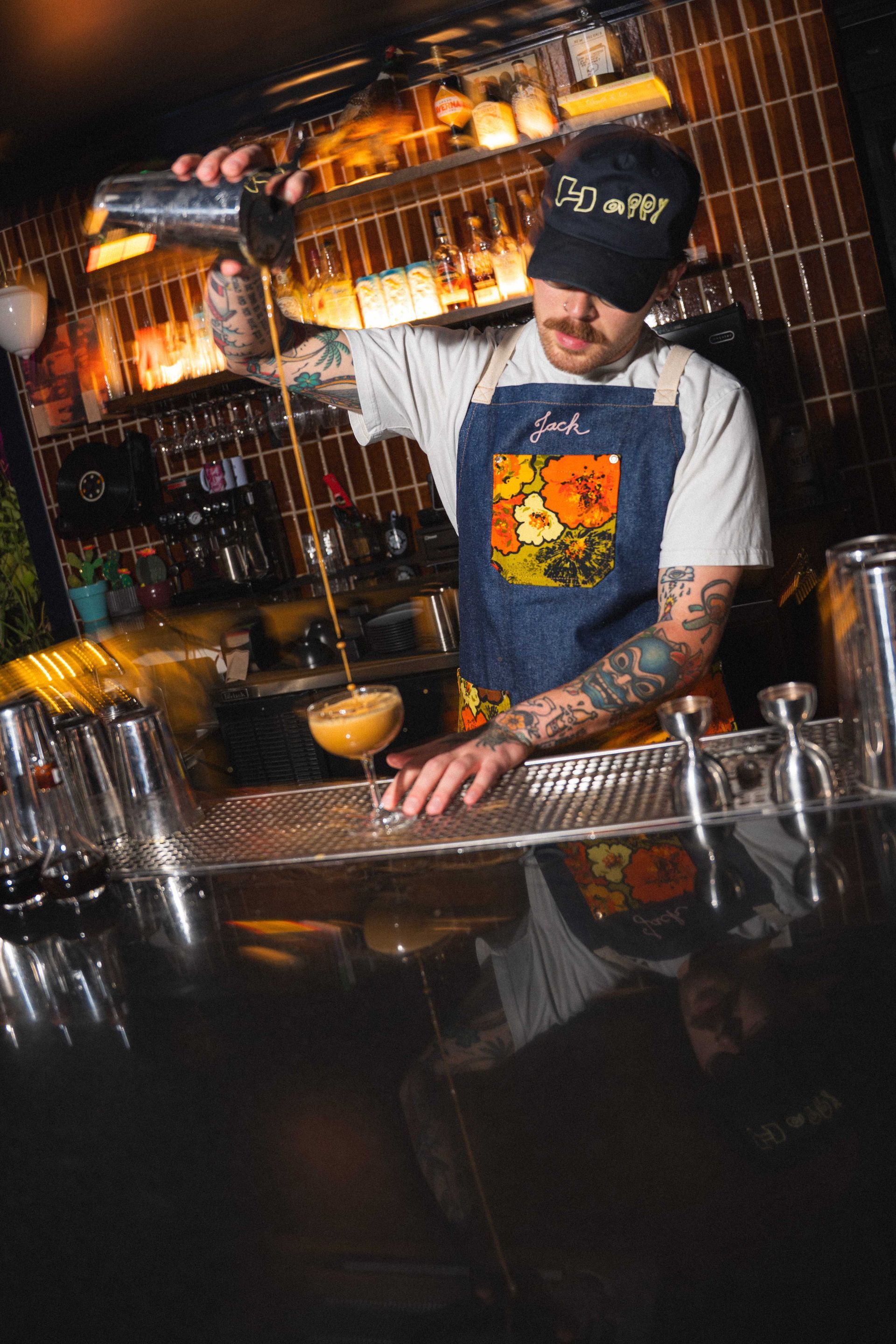 Bartender pouring a drink behind a bar, with cocktail tools and warm ambient lighting.