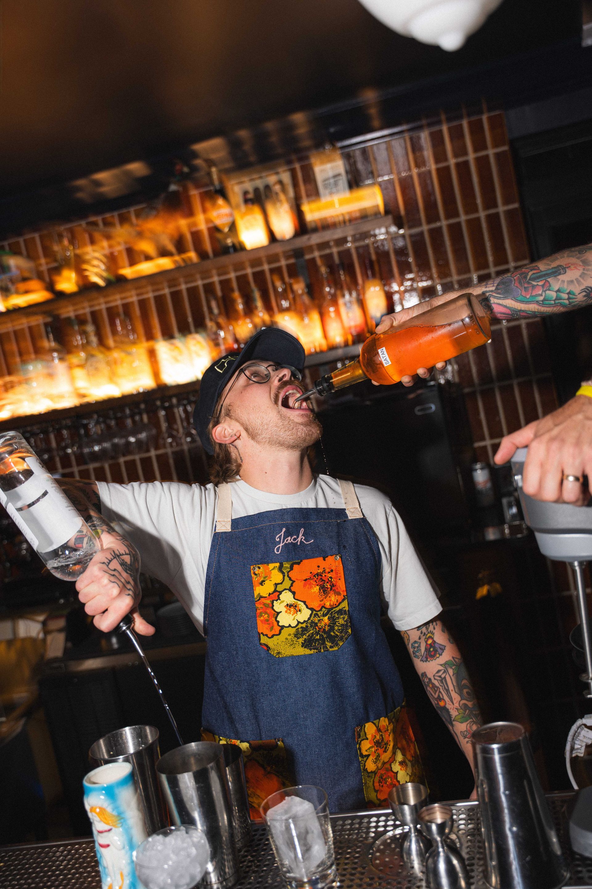 Bartender tilting head back as orange liquid pours into his mouth behind a bar with glowing shelves