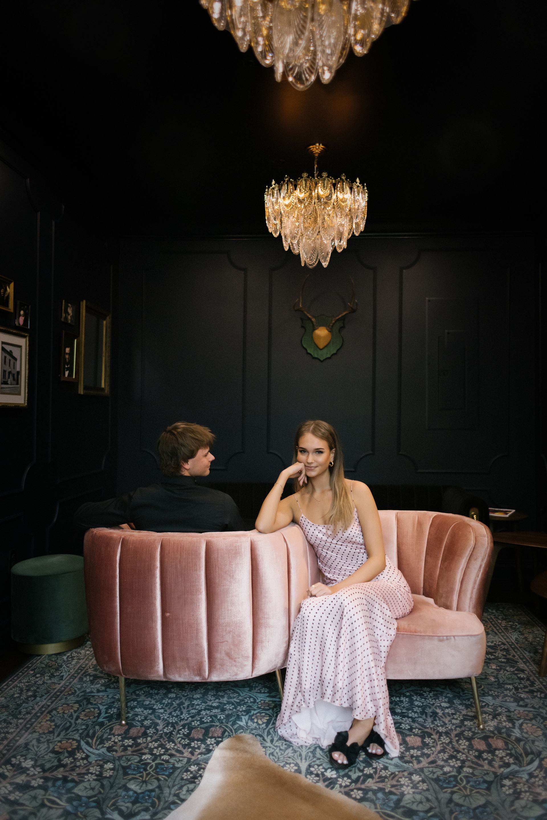 Two people seated on a pink velvet sofa in a dark, elegant lounge with chandeliers.