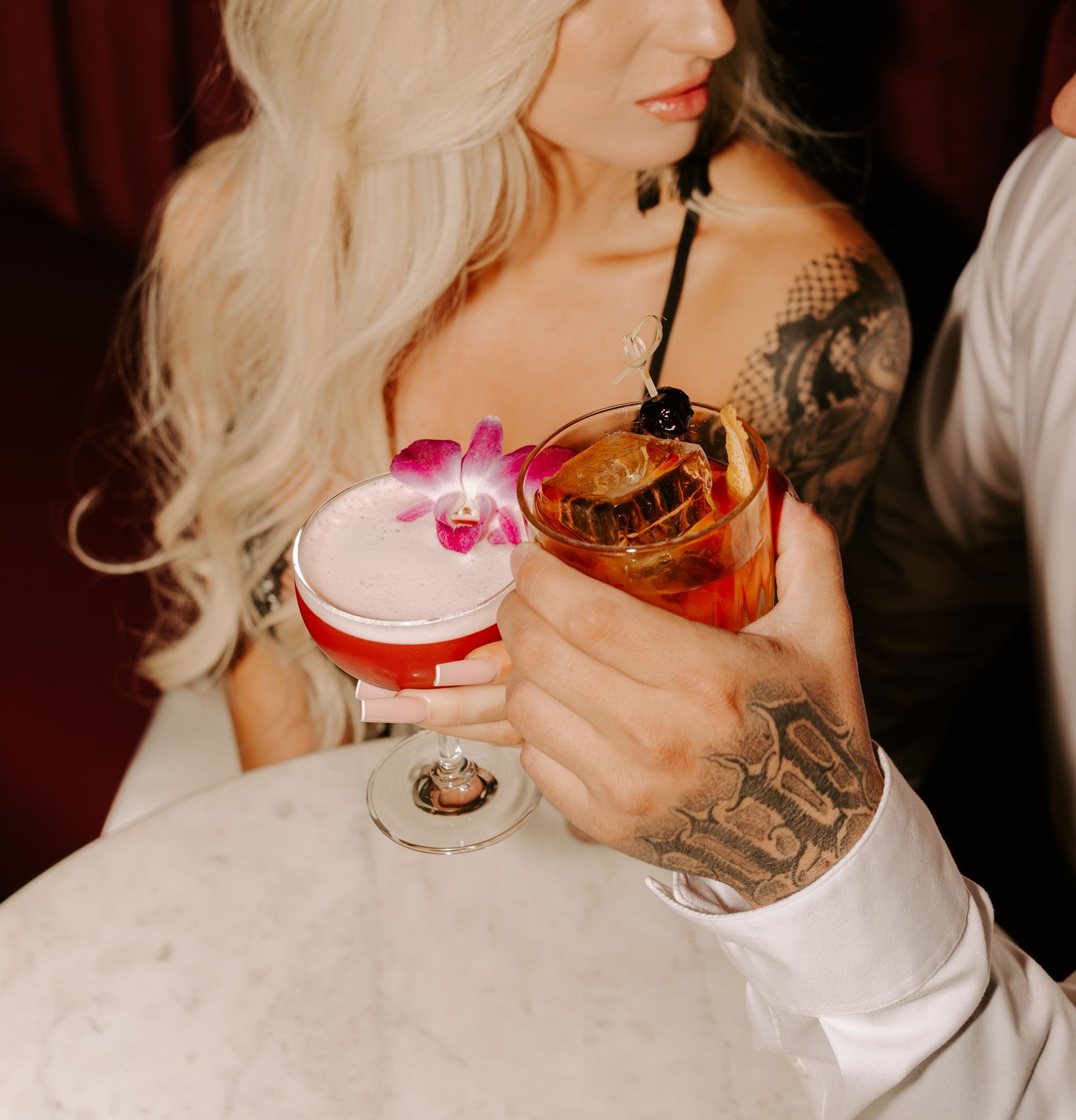 Person holding two colorful cocktails at a table, with a white rose on a pink drink and a tattooed hand visible