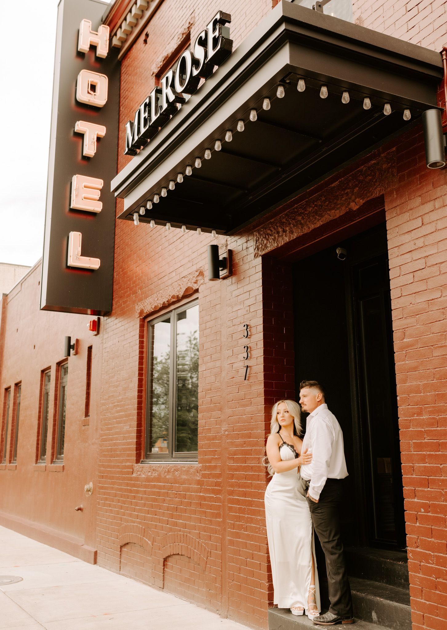 Couple standing in a hotel doorway beneath a vertical hotel sign on a brick building