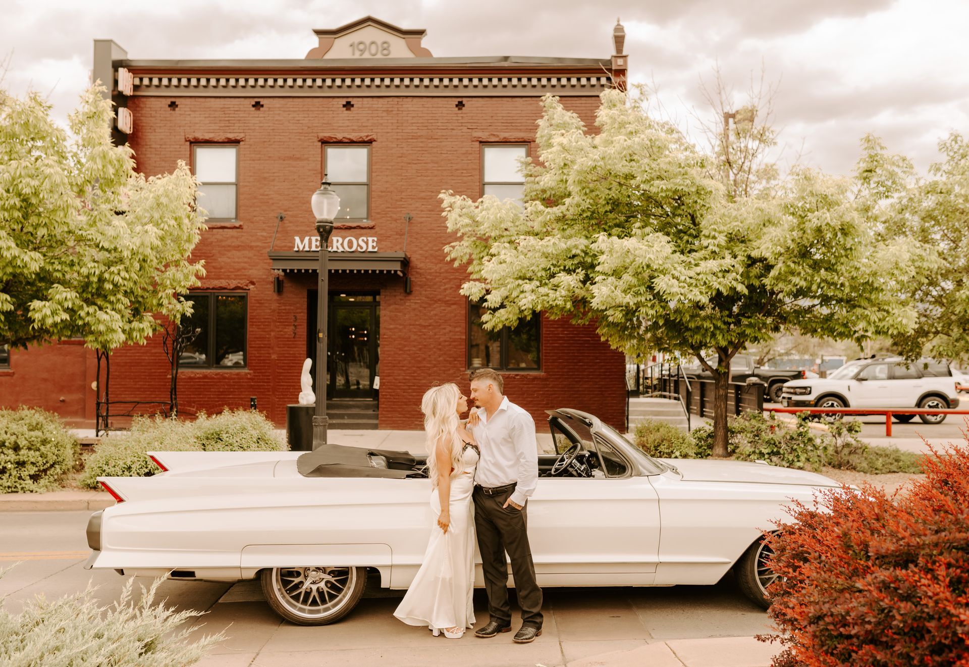 Bride and groom posing beside a white classic car in front of a red brick building