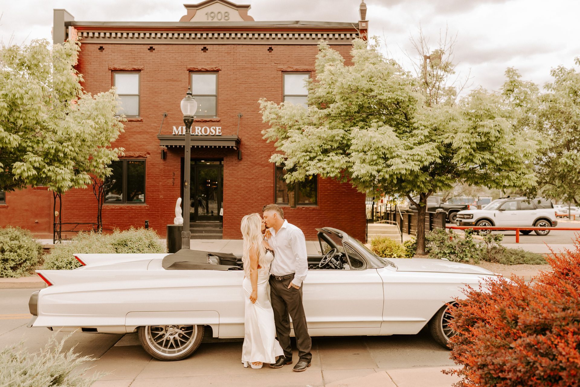 Couple standing by a white vintage car in front of a brick building and flowering trees