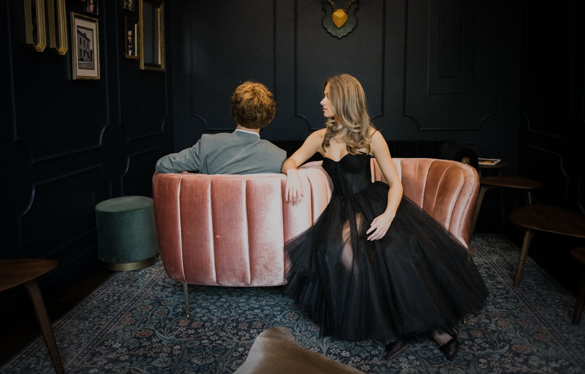 Two people seated on a pink velvet sofa in a dark, elegant room; one woman in a black dress leans toward the other.