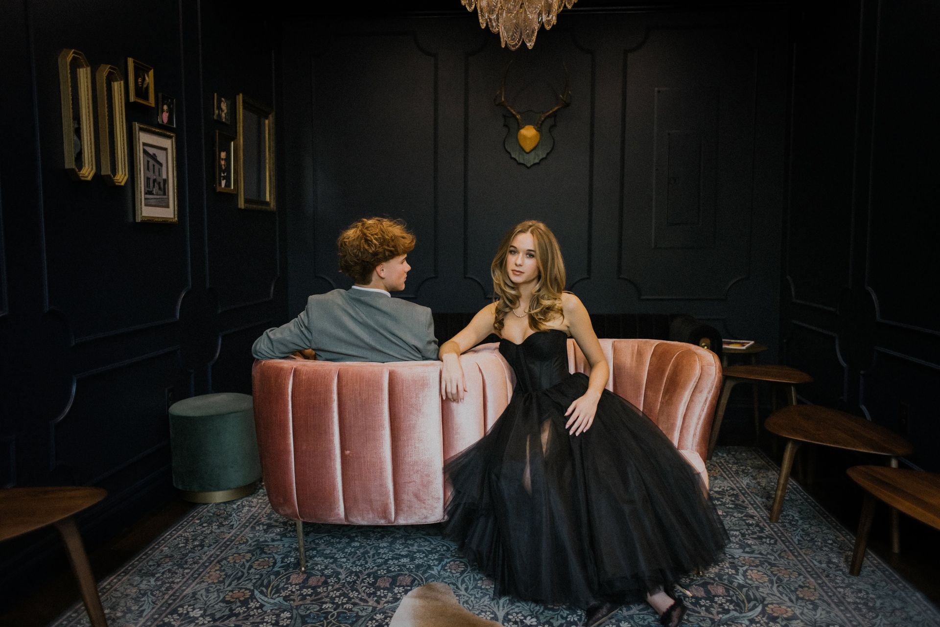 Two people seated on a pink velvet sofa in a dark, elegant room with black walls and vintage decor