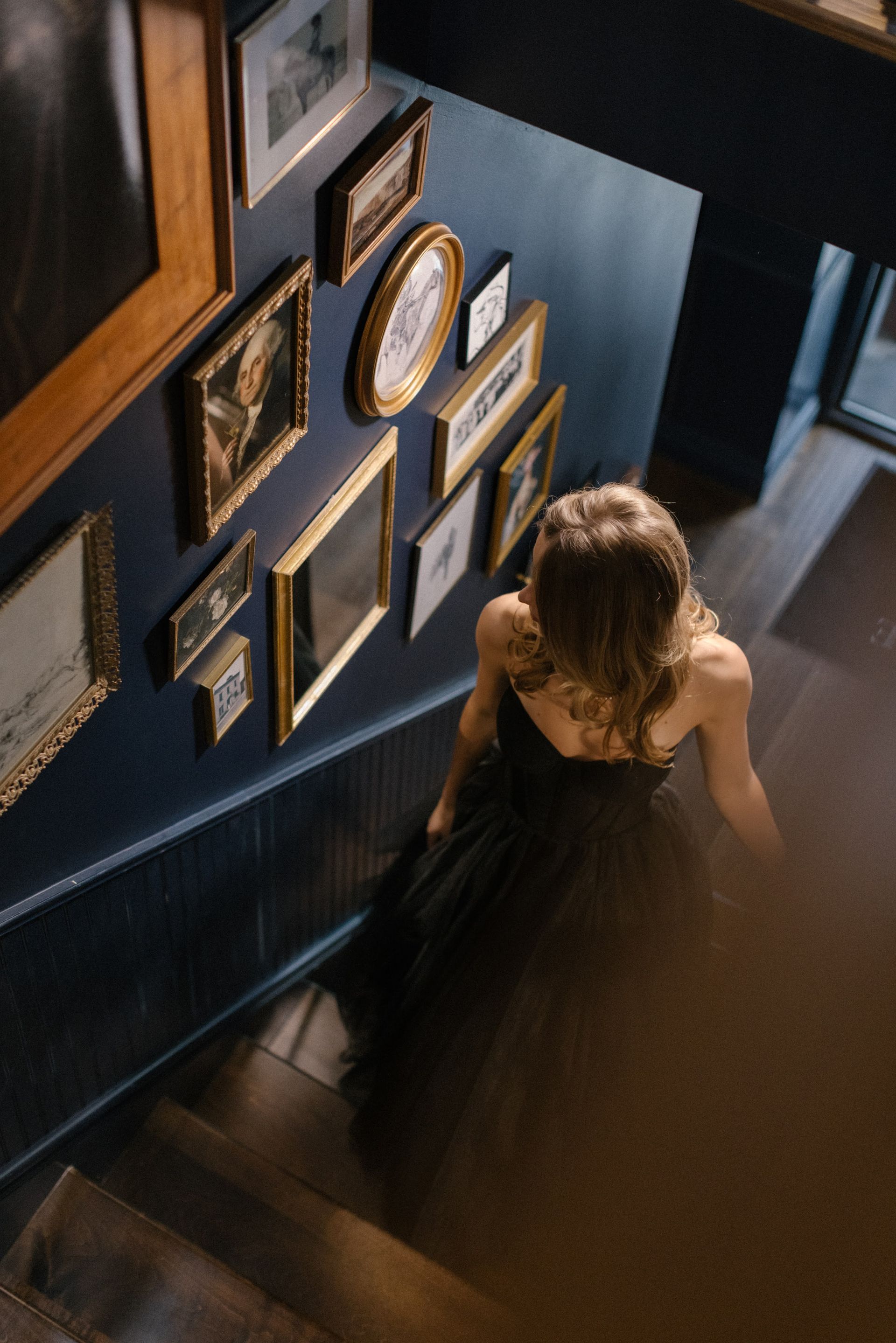 Woman in a black dress descending a dim stairway lined with framed photos