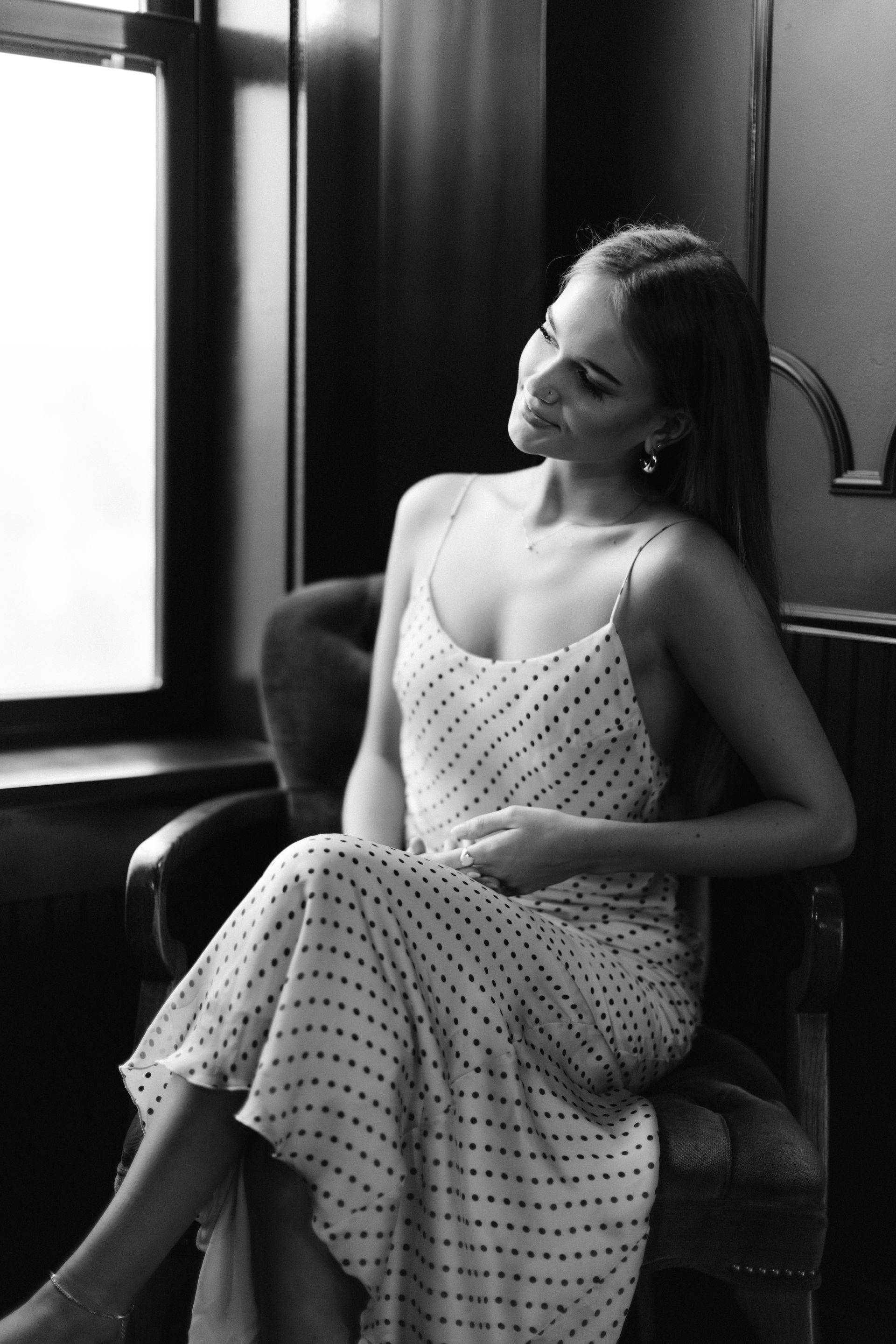 Woman in a polka-dot dress sitting in an armchair by a window, smiling softly.