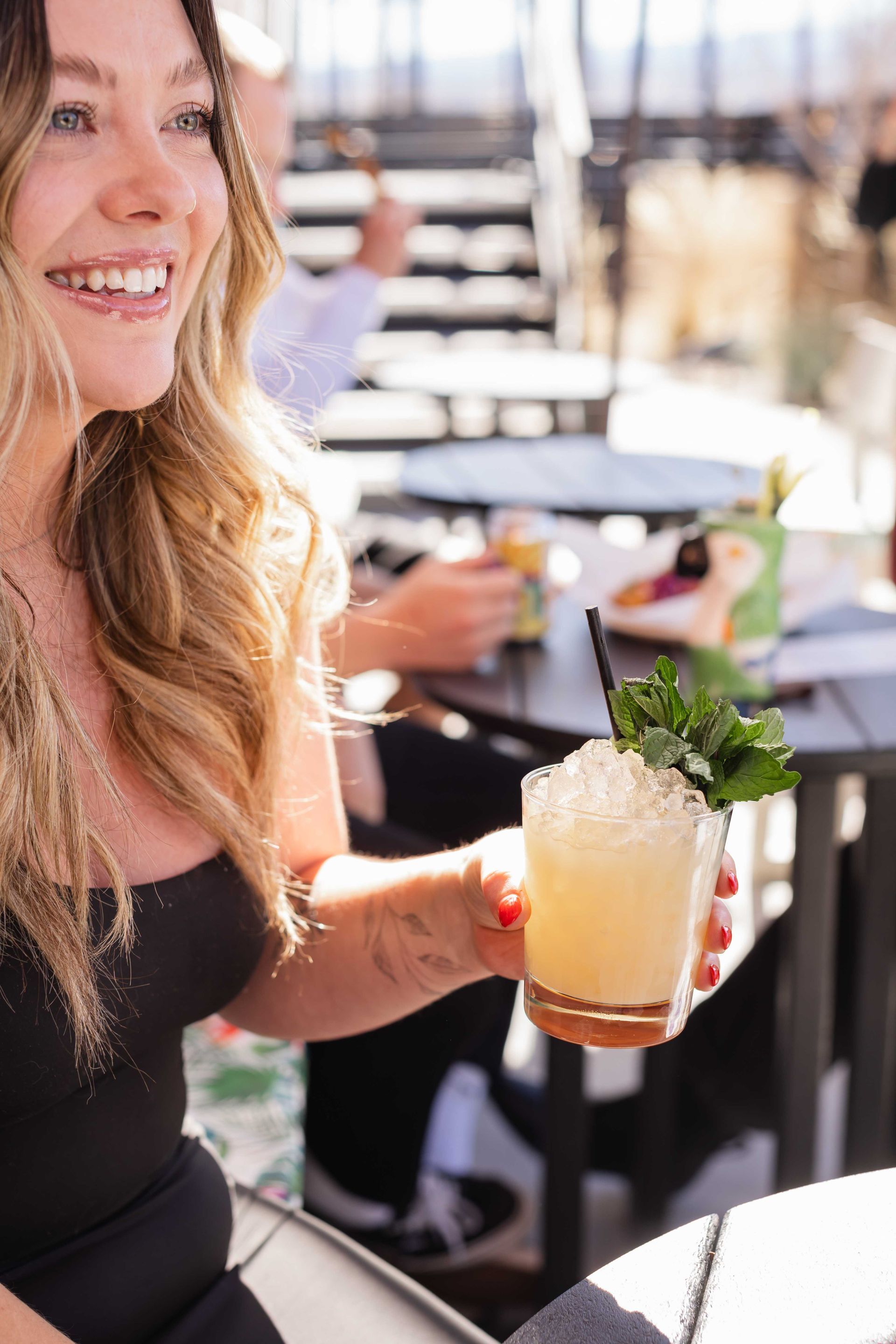 Woman holding a pale yellow cocktail with mint in a bright outdoor café seating area