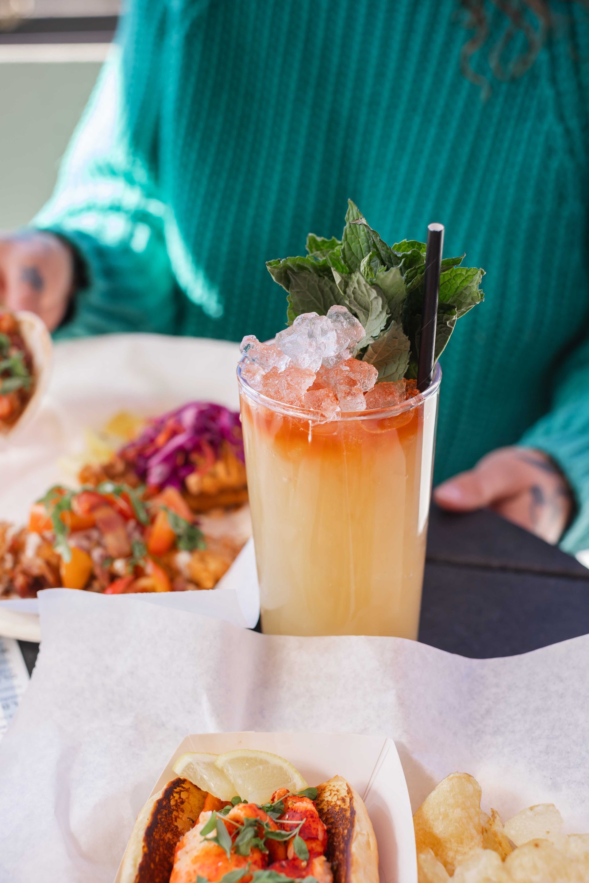 Cocktail with mint and ice on a table beside tacos and chips, with a person in a teal sweater behind it
