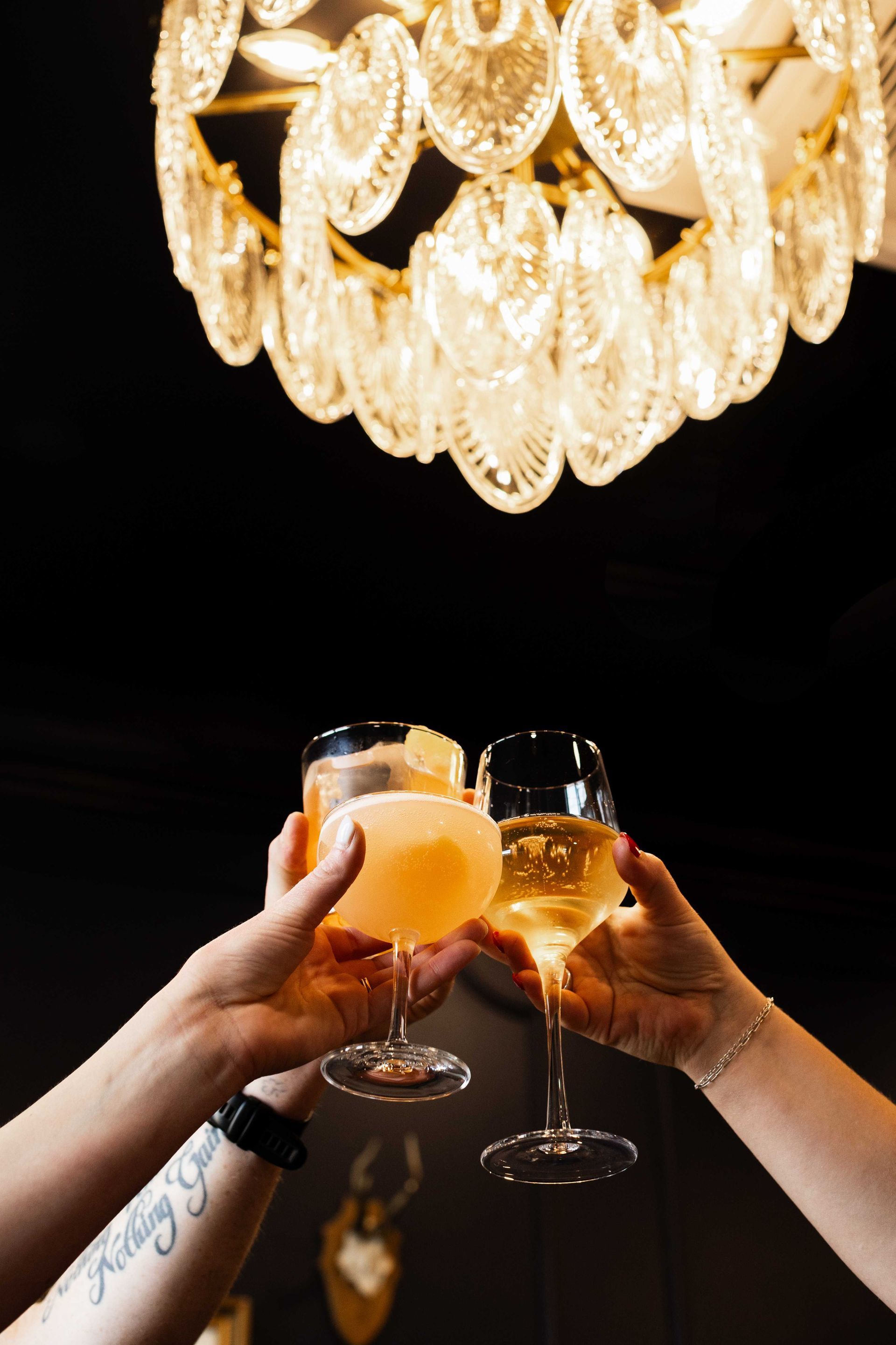 Hands toasting glasses under a glowing chandelier at a dimly lit celebration