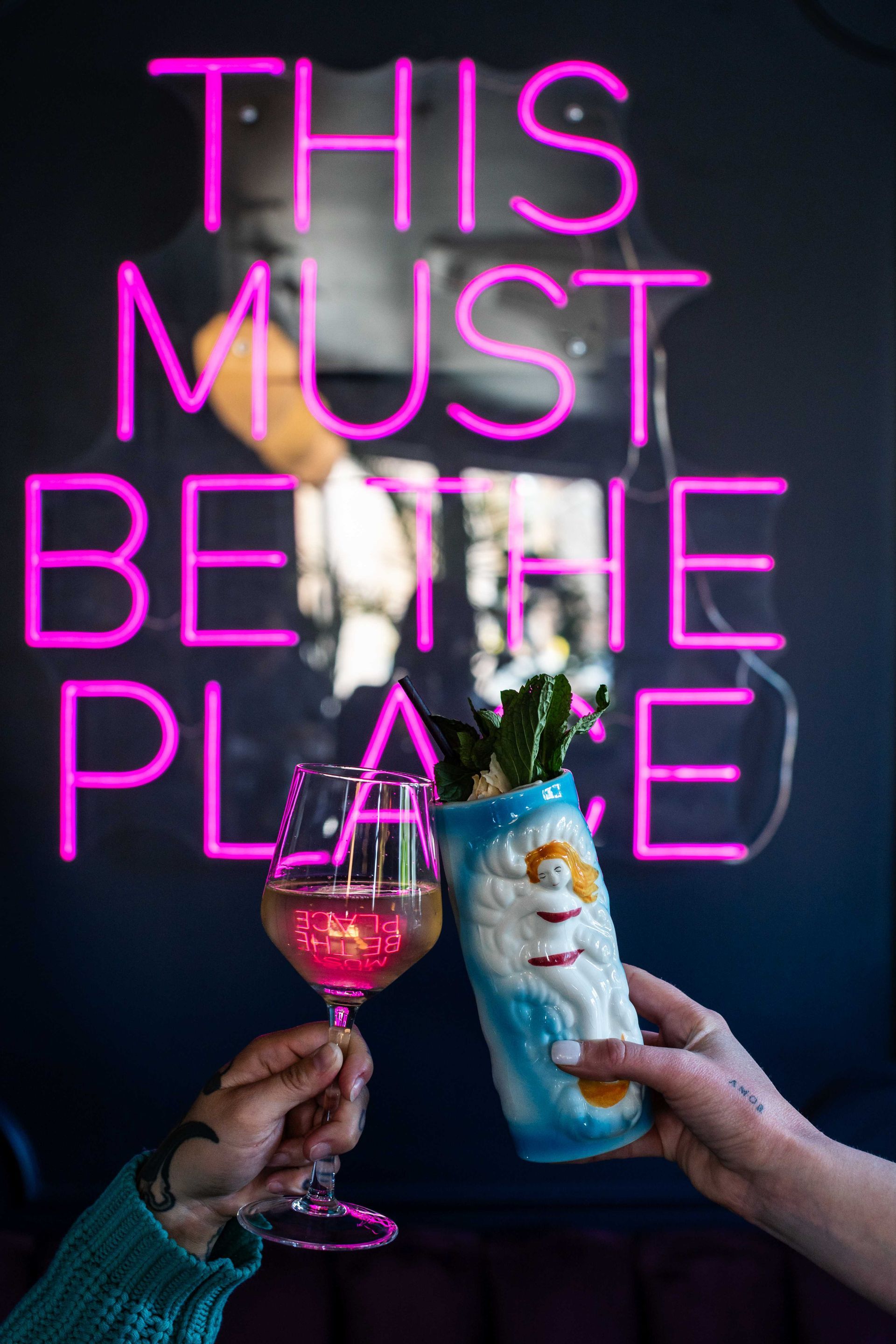 Two hands toasting champagne and a wine glass under neon pink text reading “THIS MUST BE THE PLACE.”