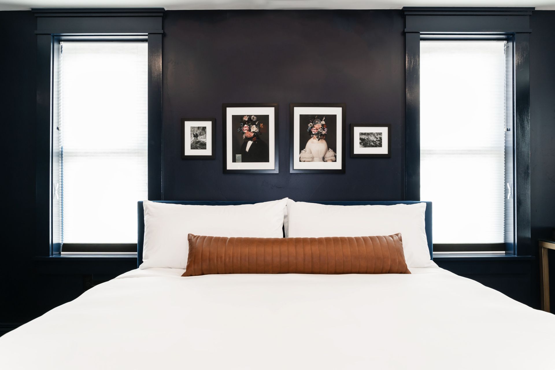 Modern bedroom with a white bed, brown lumbar pillow, and framed art on a dark wall