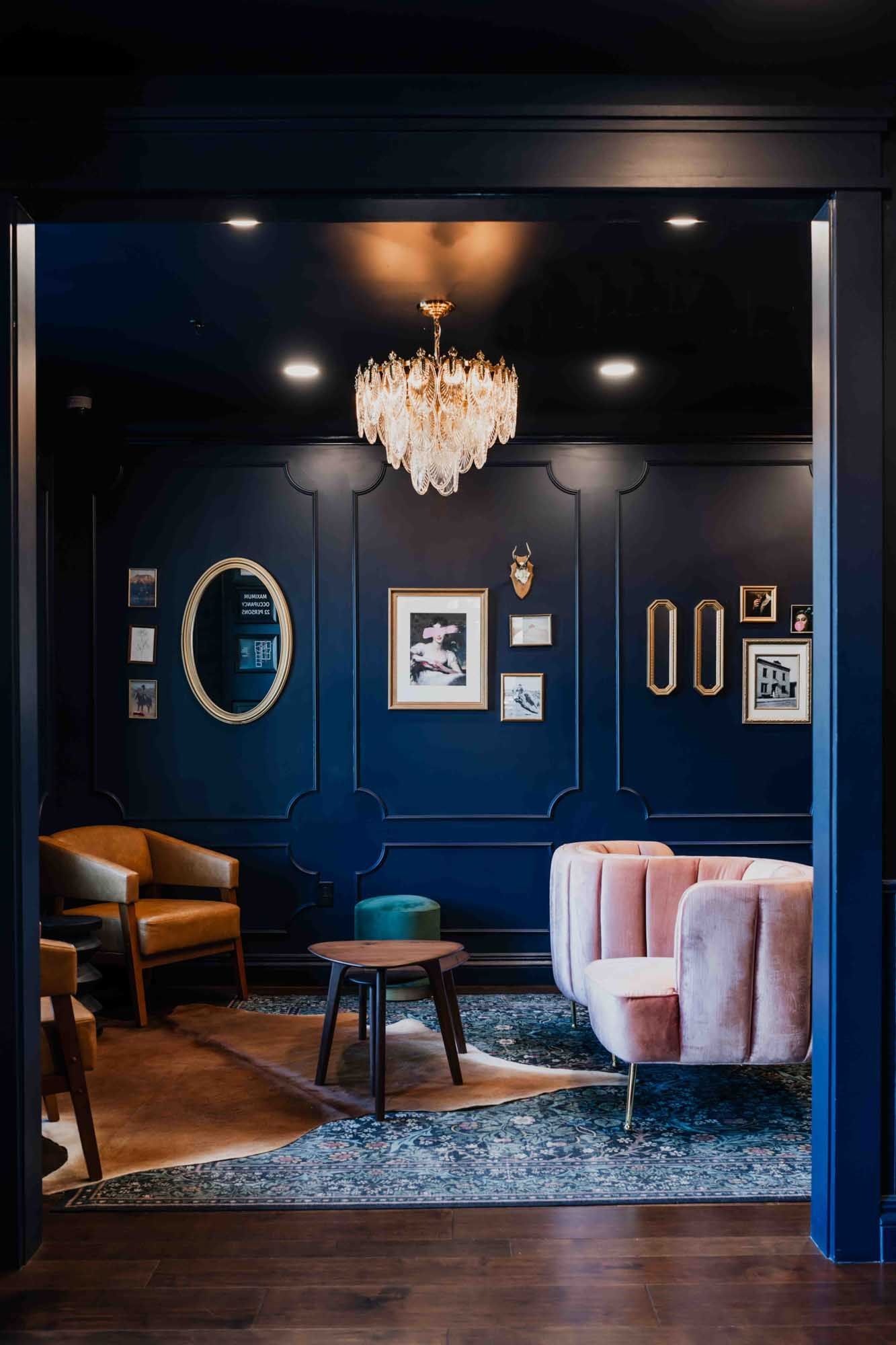 Stylish navy lounge with pink chairs, chandelier, framed art, and teal rug.