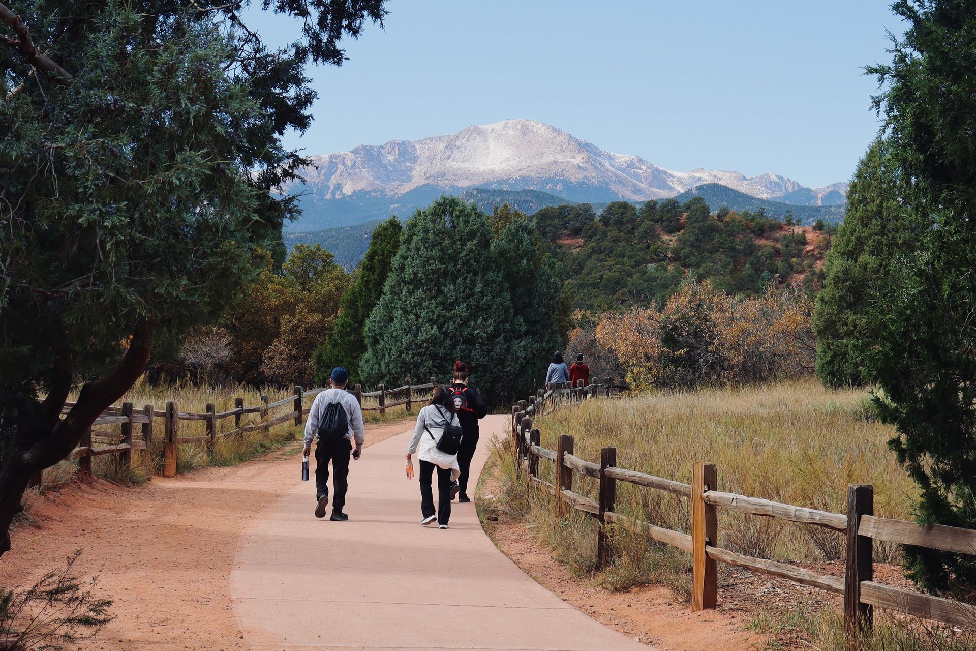 Two people walk on a dirt path beside a fence, with trees and a snow-capped mountain in the background.